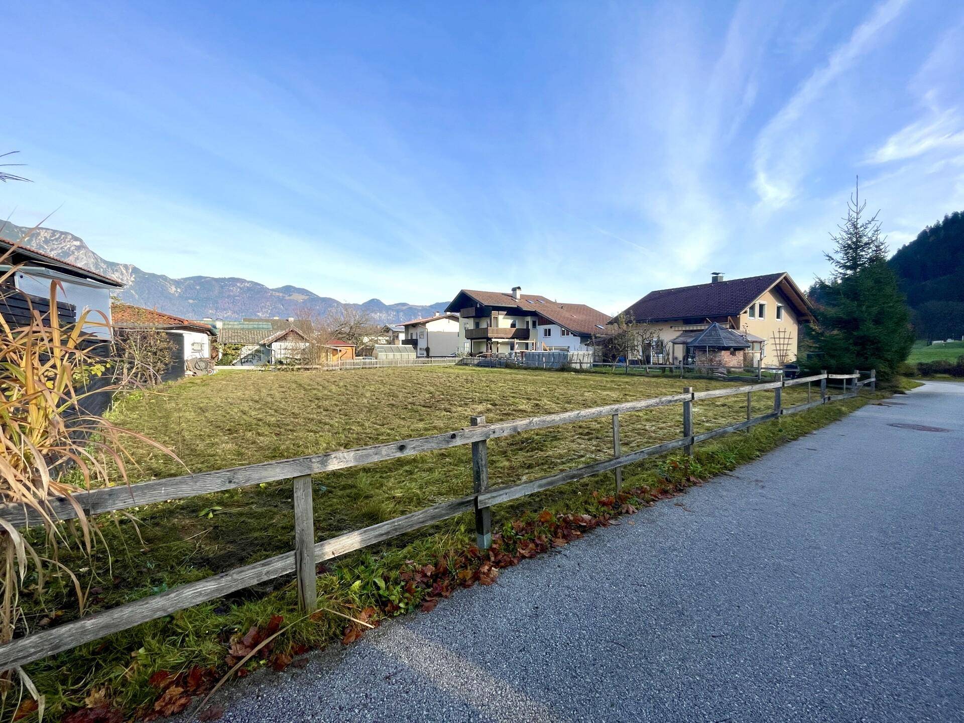 Offenes Grundstück mit Grasfläche, umgeben von traditionellen Häusern und einer Berglandschaft.
