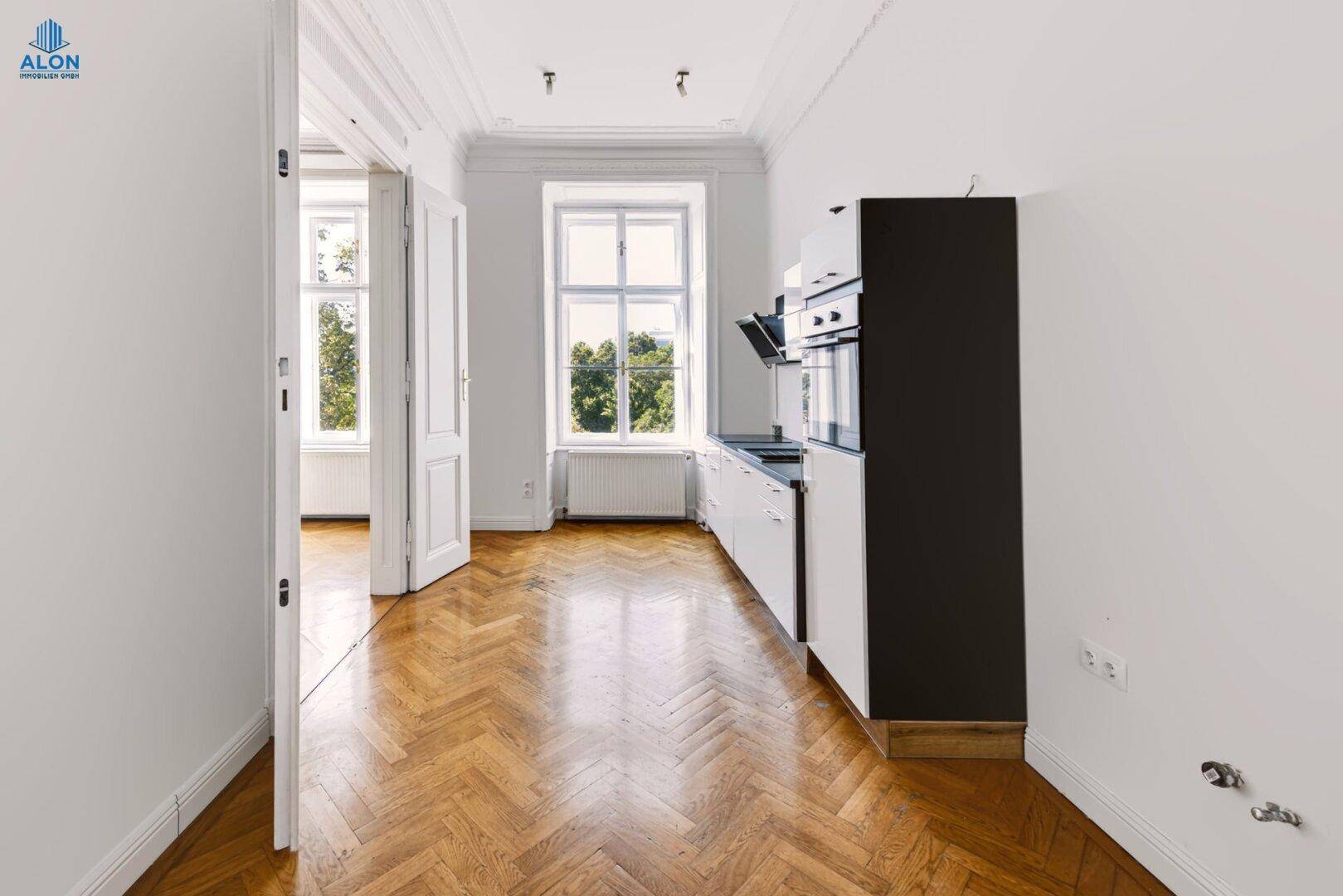 Schmale Küche mit weißer Einbauküche, Fischgrätparkett und Fenster mit Grünblick, gut beleuchtet.