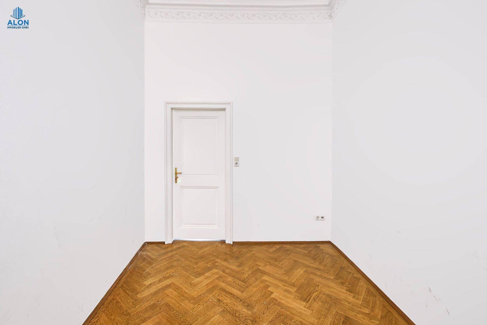 Schlichtes Schlafzimmer mit Fischgrätparkett, hohen Decken und weißer Tür, in gutem Zustand.
