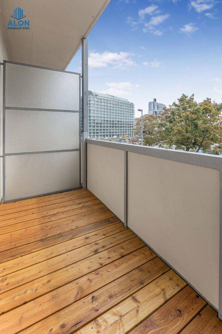 Sonniger Balkon mit Holzdielen und seitlichem Blick auf die Stadt, umgeben von Grünflächen.