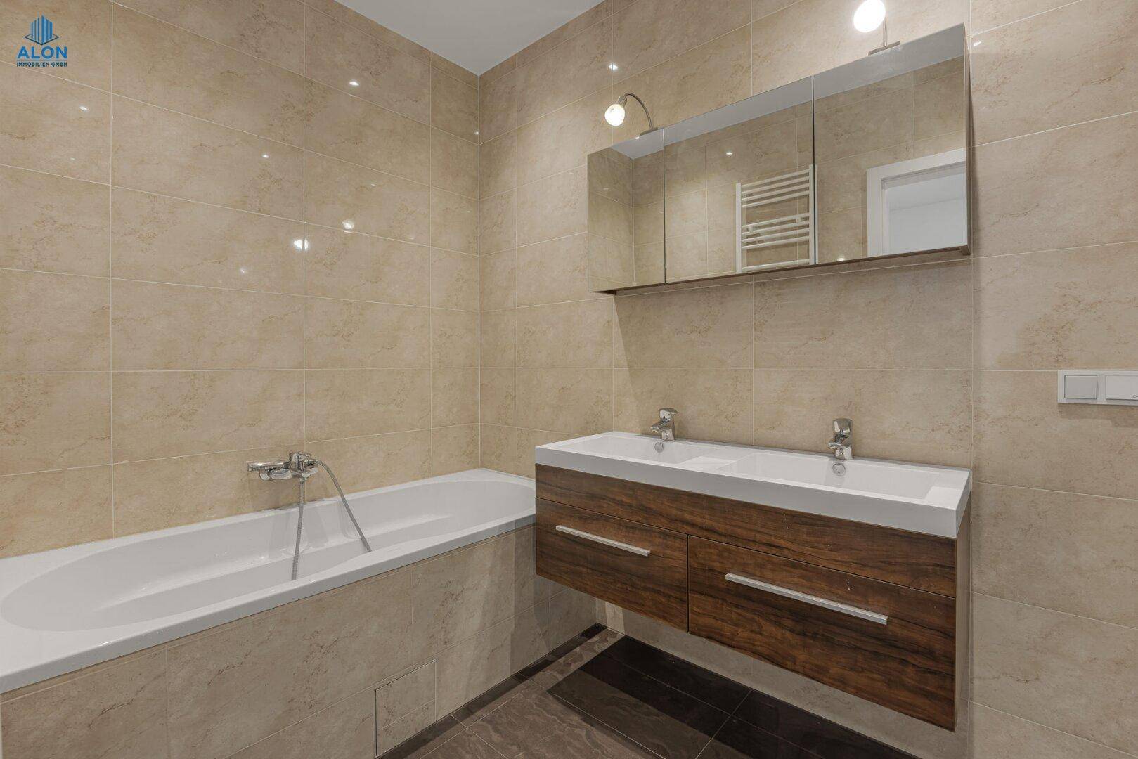 Modernes Badezimmer mit Badewanne, Doppelwaschbecken und beigefarbenen Fliesen, inklusive Spiegelschrank.