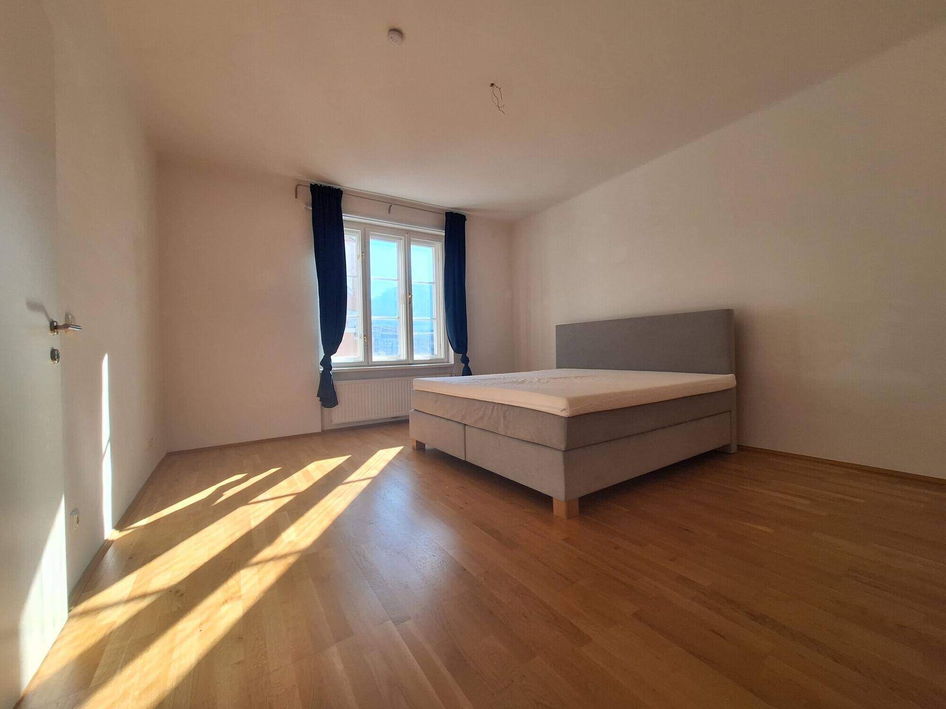 Schlafzimmer mit Parkettboden, einem Doppelbett und blauen Vorhängen, sonnendurchflutet.