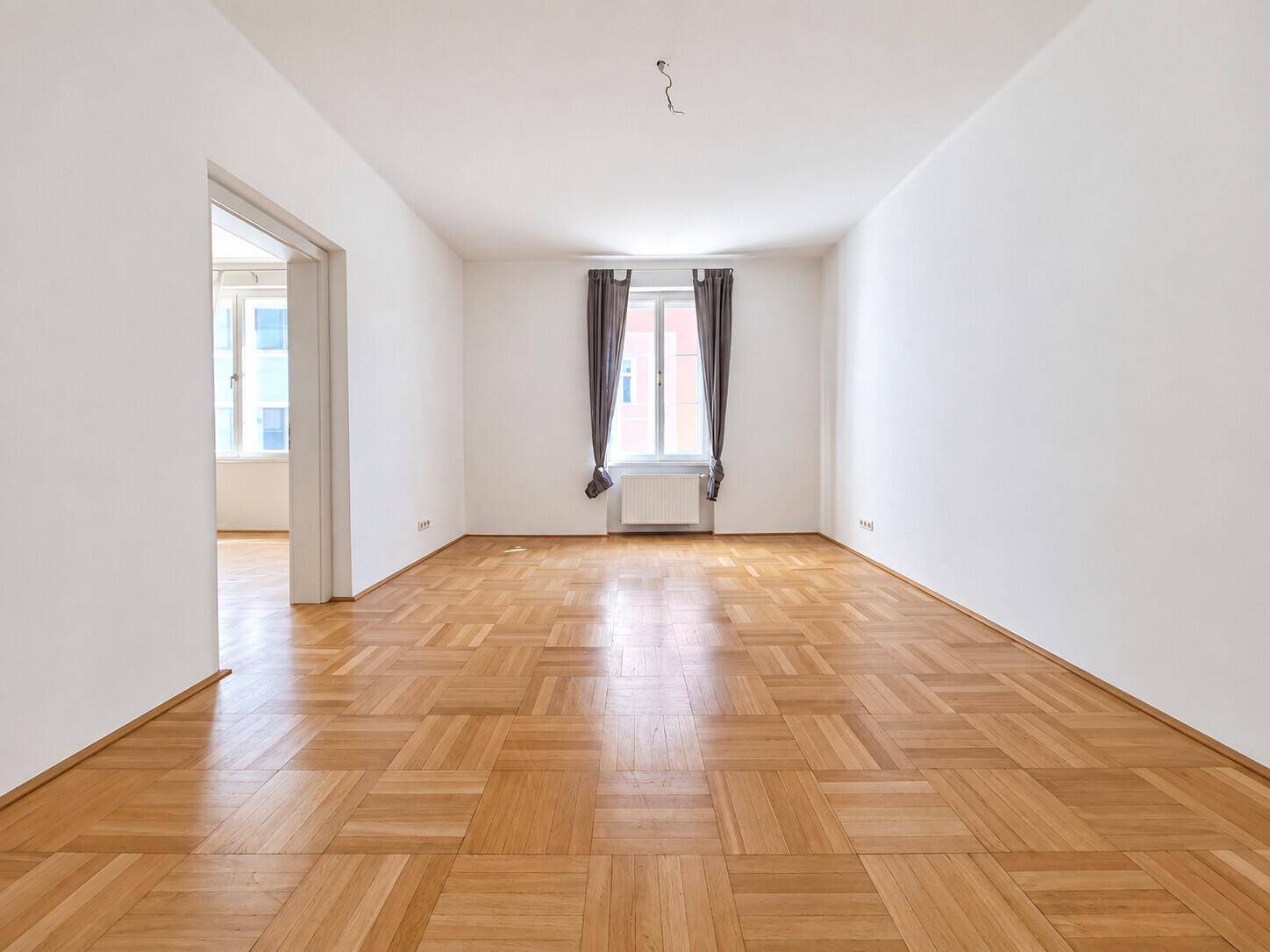Geräumiges Wohnzimmer mit Parkettboden und einem Fenster mit dunklen Vorhängen, unmöbliert.