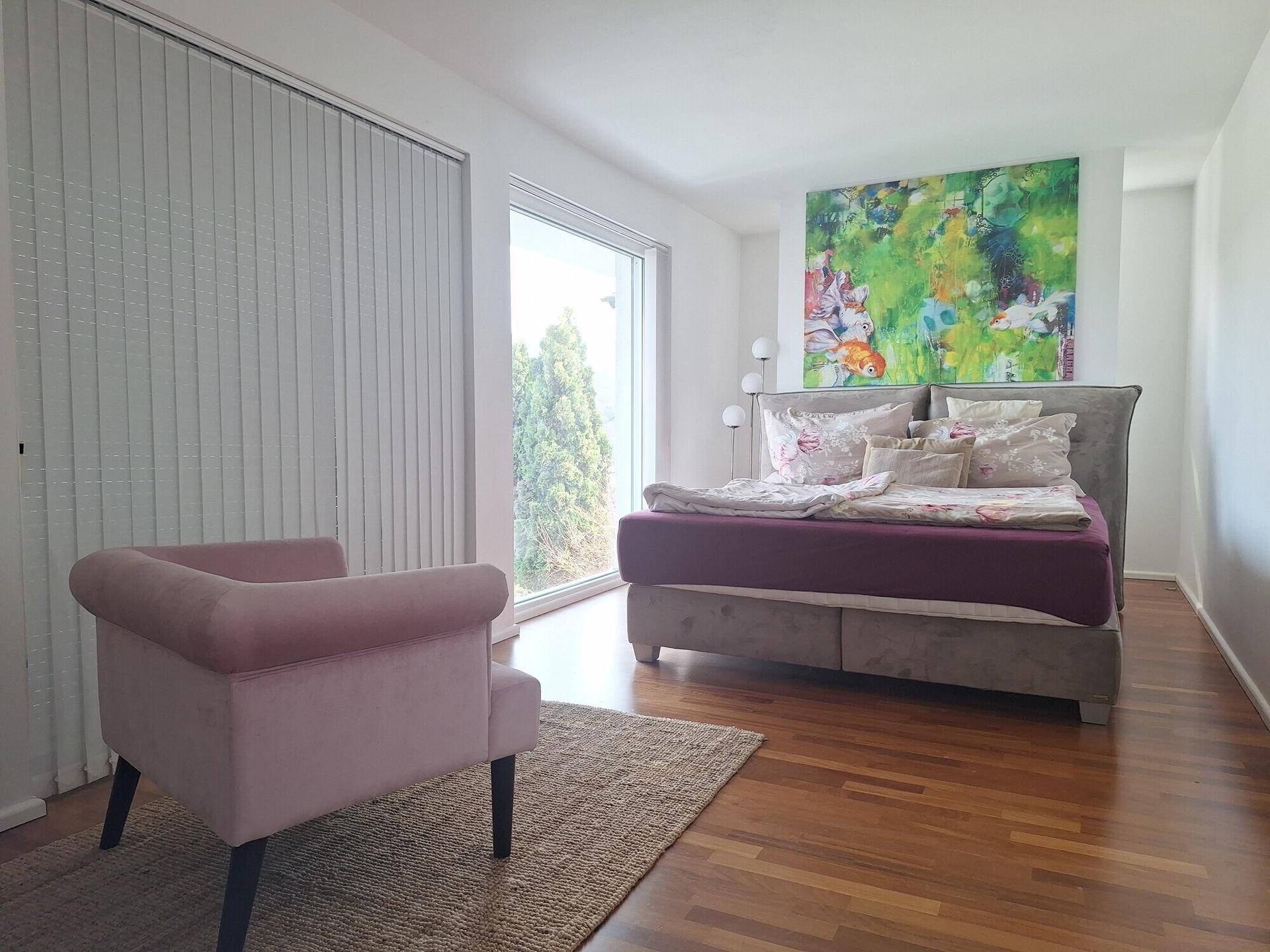 Helles Schlafzimmer mit Holzboden, großem Fenster und modernem Kunstwerk an der Wand.