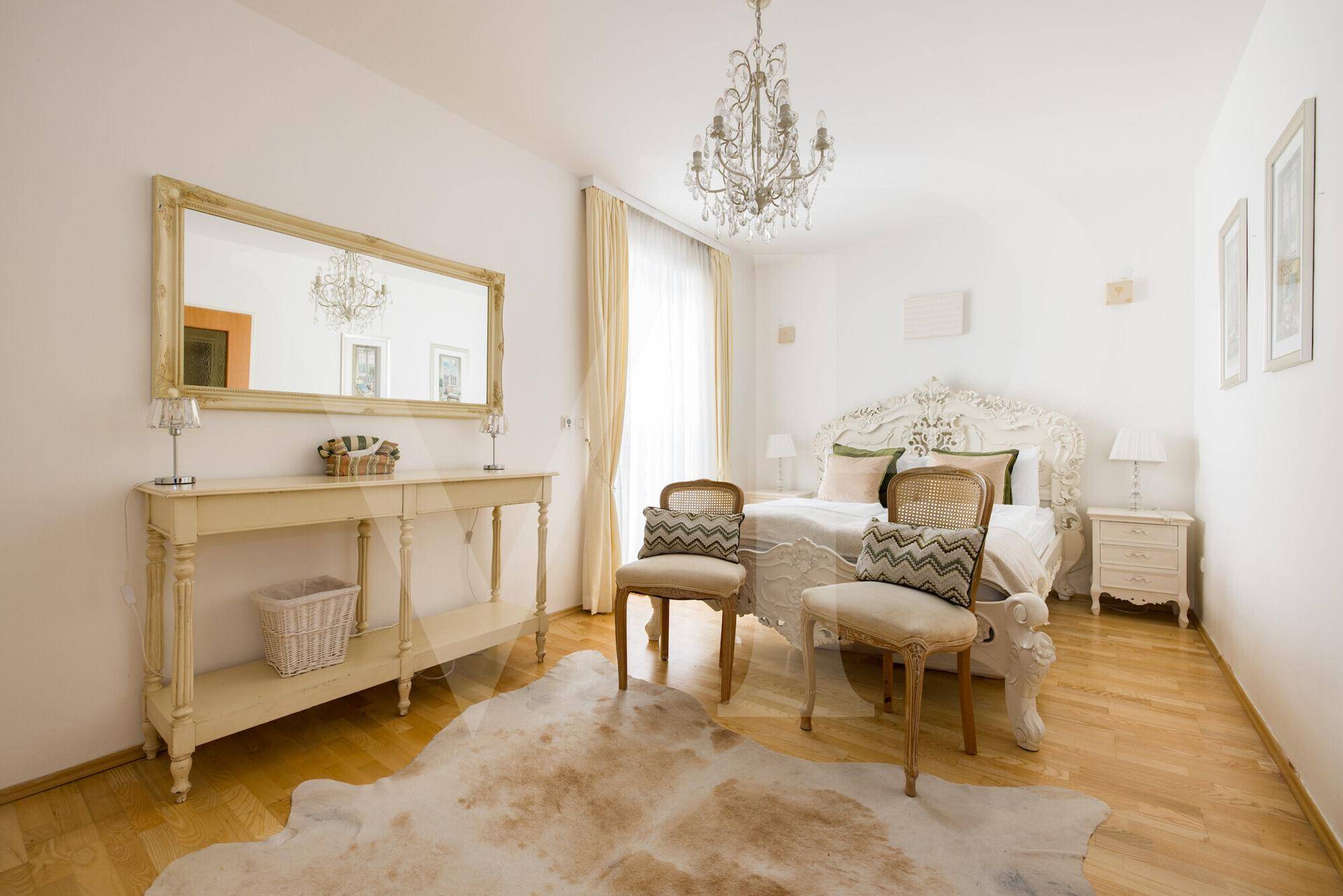 Elegantes Schlafzimmer mit weißem Bett, großem Spiegel und Holzboden, beleuchtet von einem Kronleuchter.