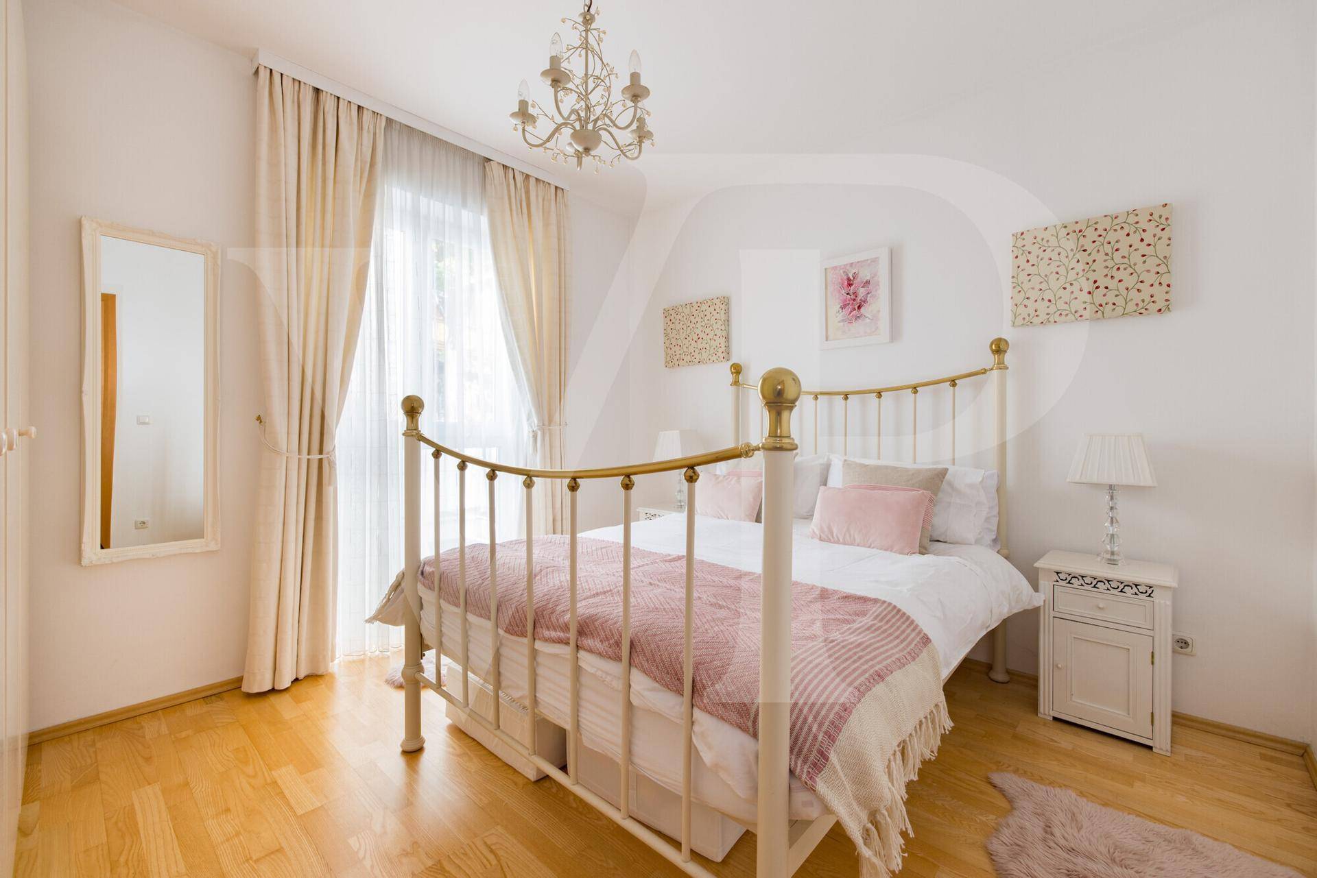 Helles Schlafzimmer mit einem eleganten weißen Metallbett, Holzboden und großen Fenstern mit Vorhängen.