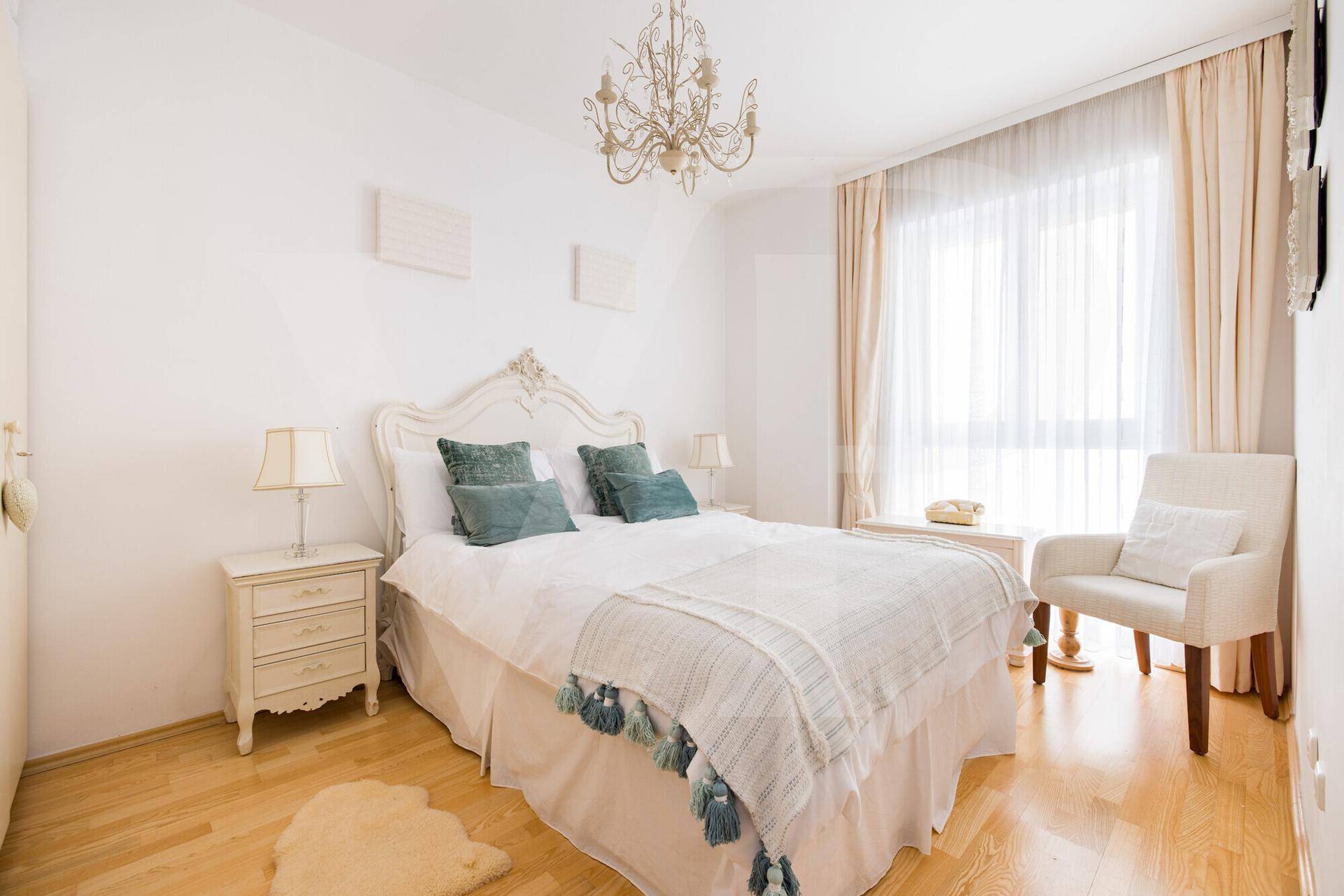 Helles Schlafzimmer mit weißem Bett, blauen Akzenten und einem Kronleuchter, der den Raum erhellt.