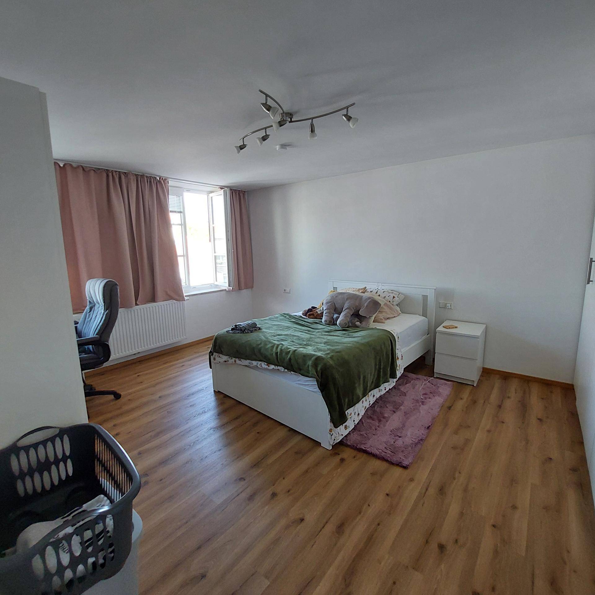 Gemütliches Schlafzimmer mit Laminatboden, großem Fenster und Platz für einen Arbeitsbereich.