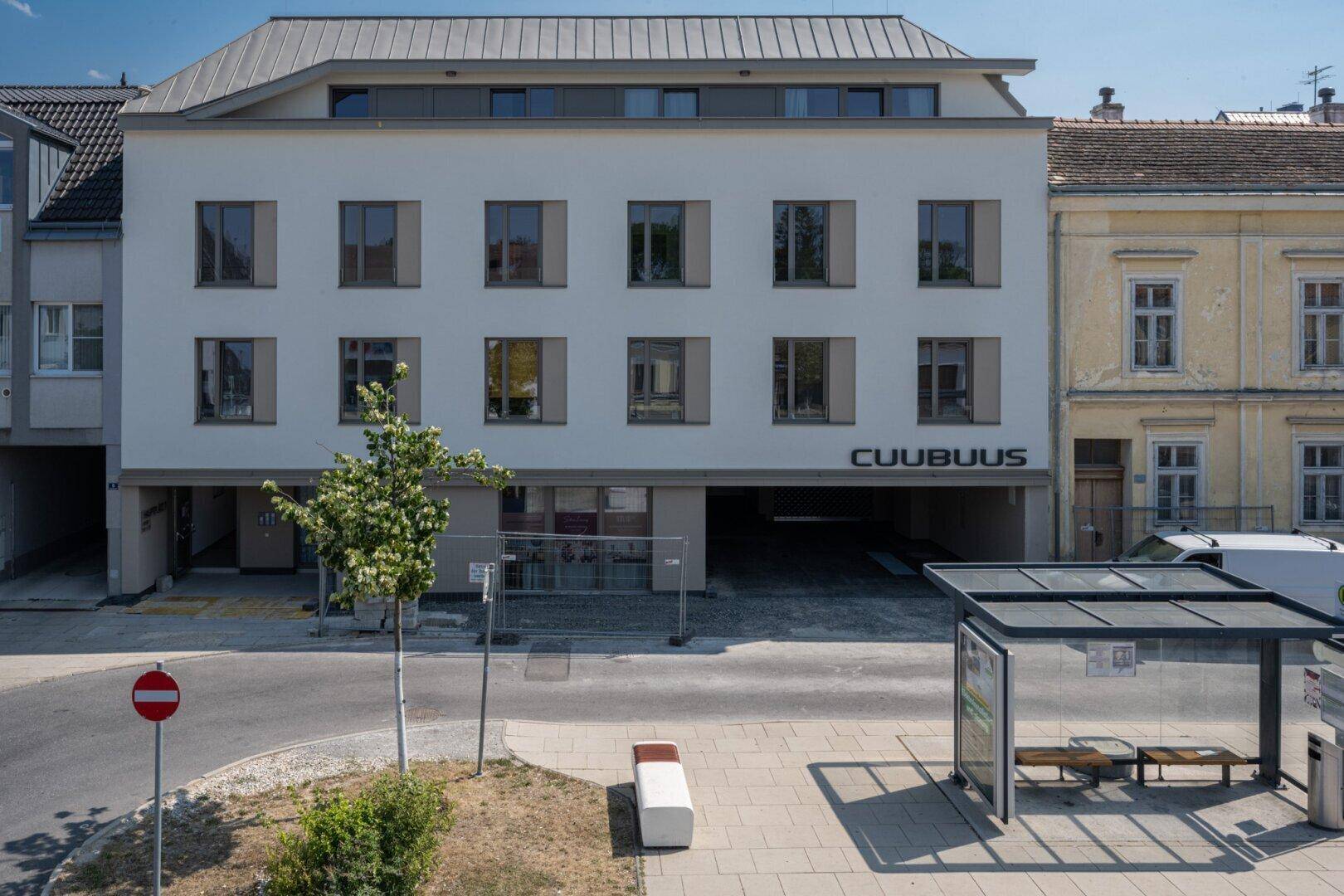 Modernes Mehrfamilienhaus mit klarer Architektur, großen Fenstern und einer Bushaltestelle im Vordergrund.
