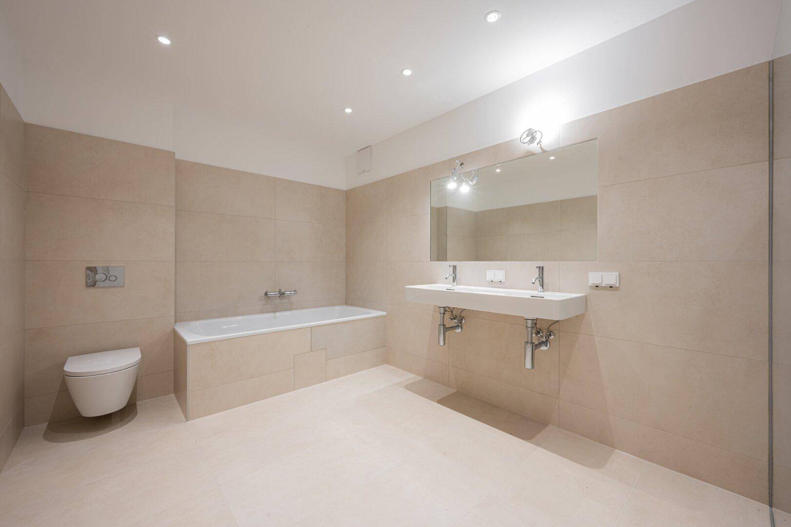 Modernes Badezimmer mit beigefarbenen Fliesen, Badewanne, Doppelwaschbecken und Wand-WC.