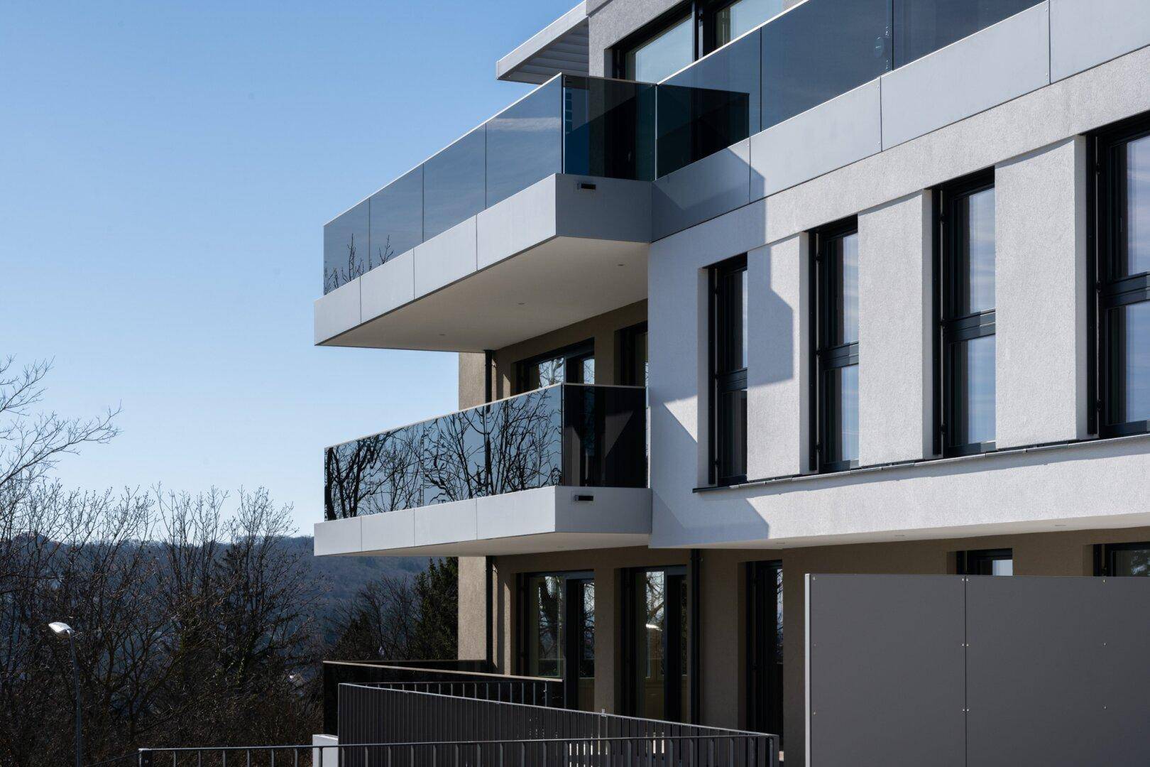 Moderne Wohngebäude mit mehreren Balkonen und Glasgeländern, umgeben von Bäumen und Hügeln.