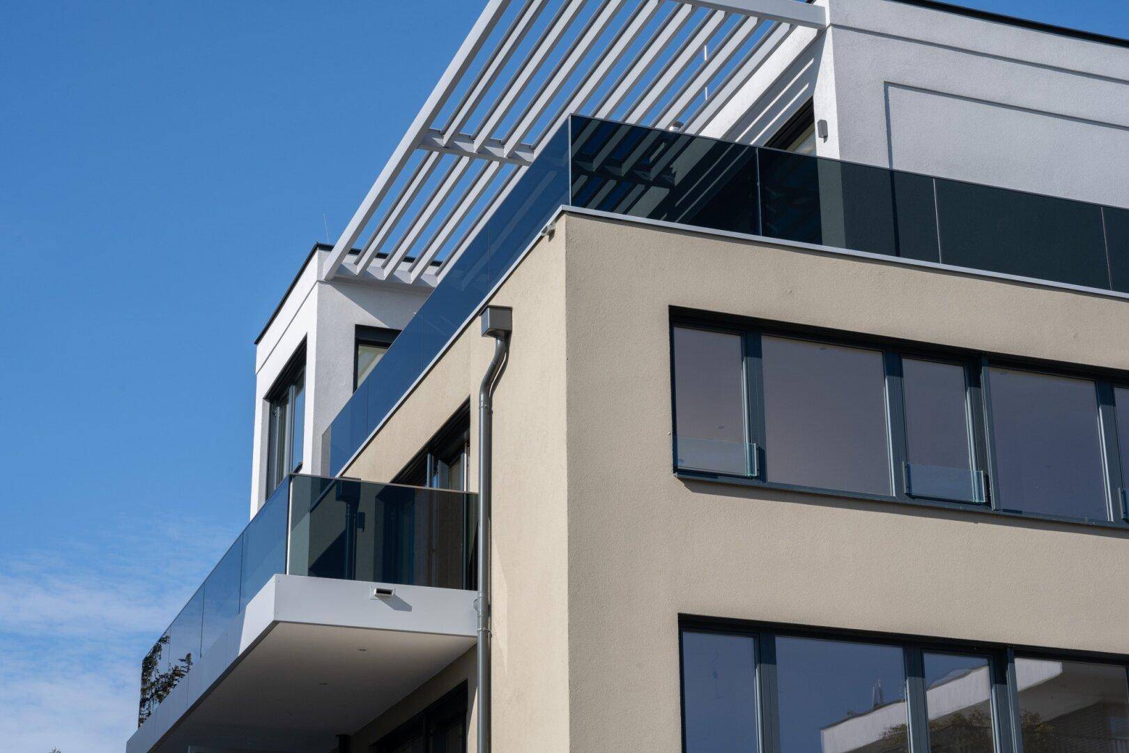 Detailansicht der modernen Fassade mit Glasbalkonen und Pergola-Struktur auf dem Dach.