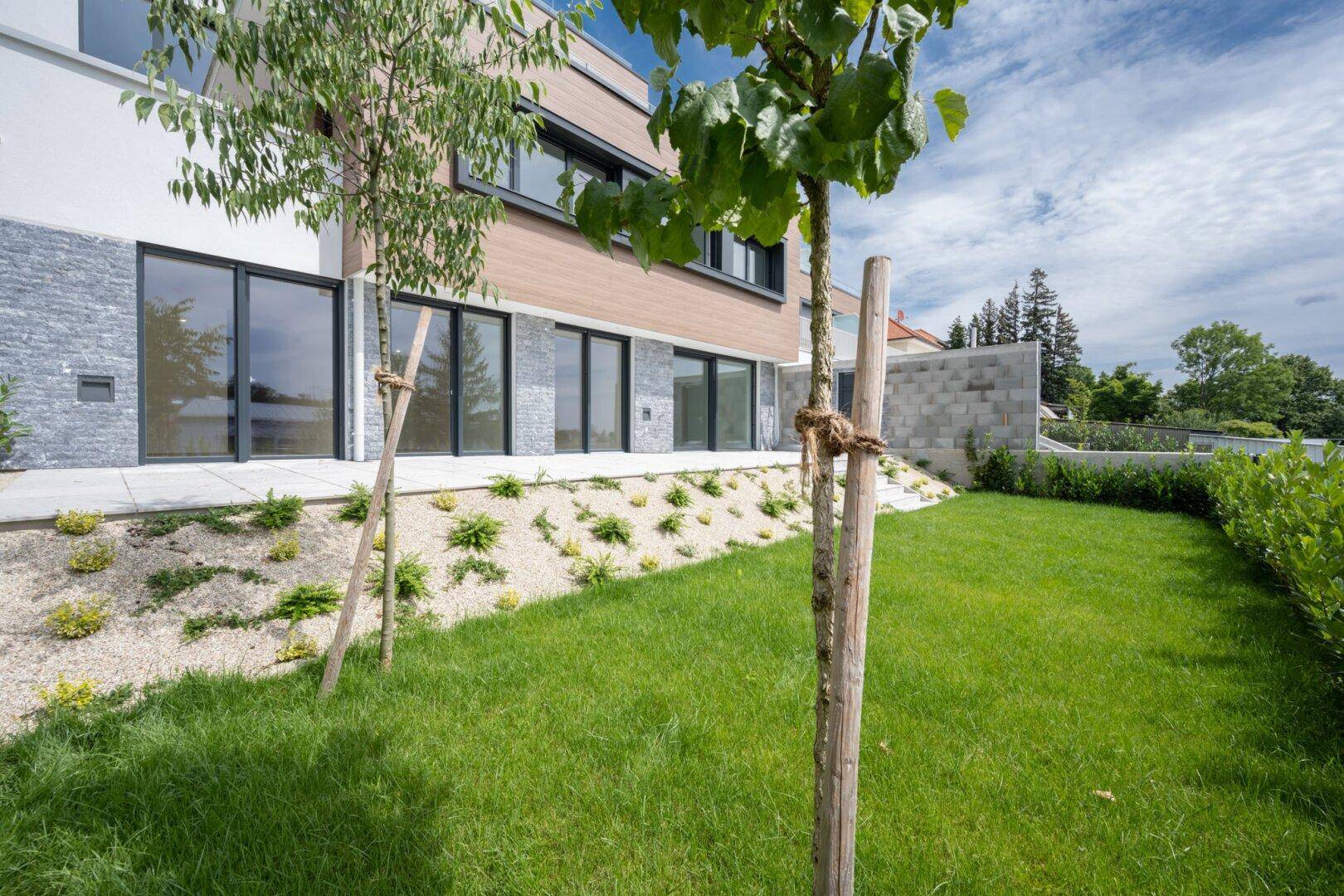 Gepflegter Gartenbereich mit Rasenfläche und einer kleinen Terrasse vor den großen Glasfronten des Gebäudes.