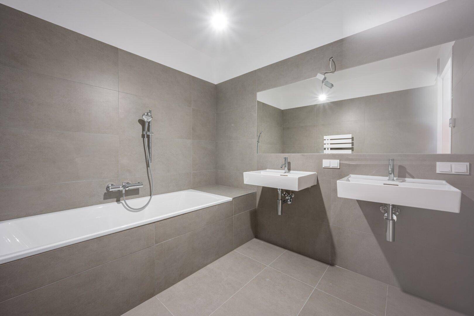Modernes Badezimmer mit grauen Fliesen, Badewanne und zwei stilvollen Waschbecken unter einem großen Spiegel.