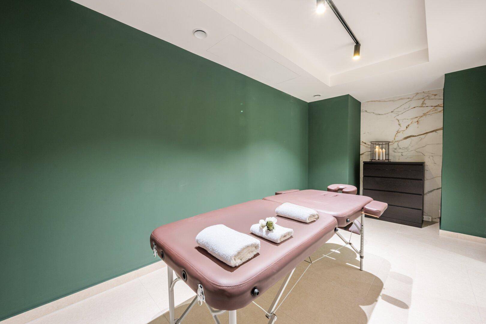 Entspannungsraum mit Massageliegen, grünen Wänden und indirekter Beleuchtung für eine ruhige Atmosphäre.
