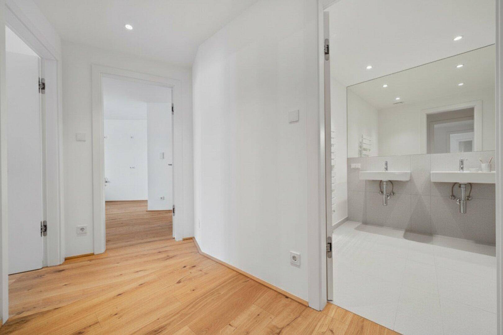 Heller Flur mit Holzboden und Zugang zu einem modernen Badezimmer mit Doppelwaschbecken.