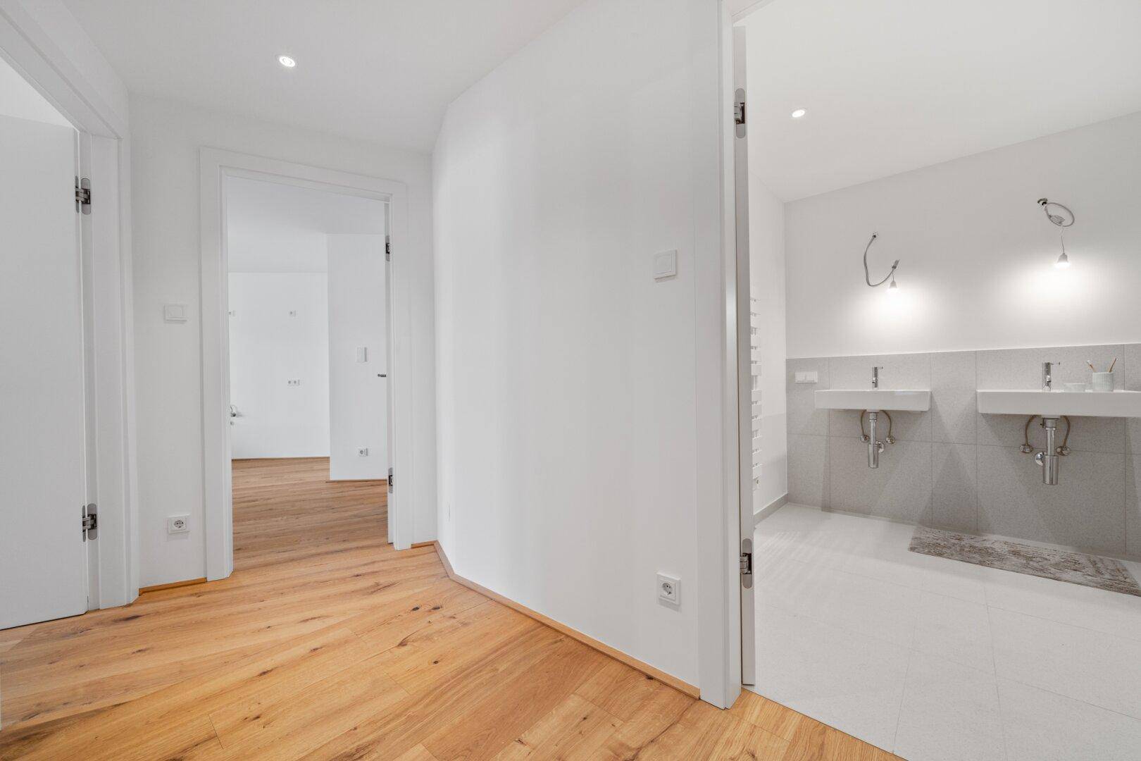 Heller Flur mit Holzboden, der zu einem Badezimmer mit zwei Waschbecken führt.
