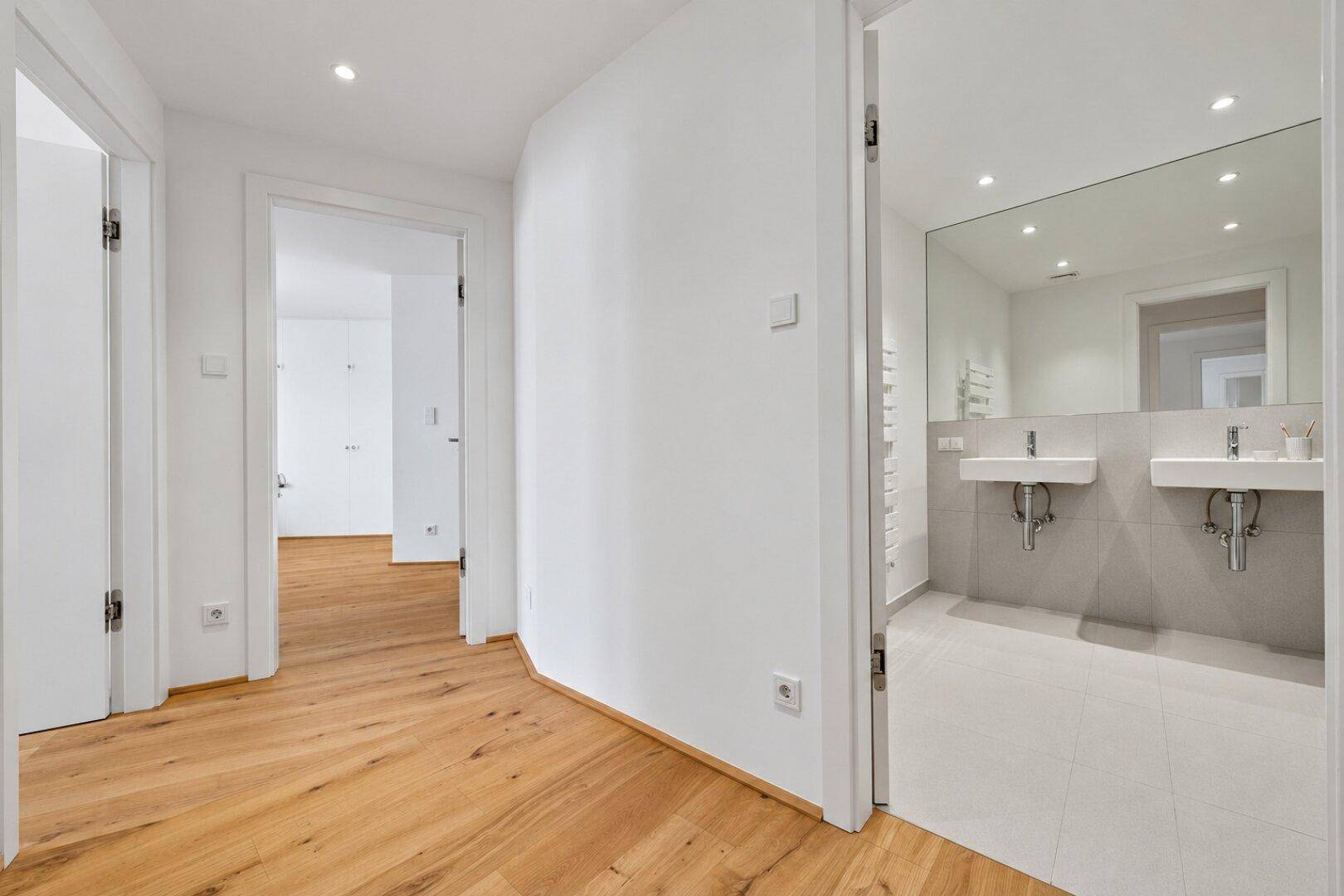 Heller Flur mit Holzboden und Zugang zu einem modernen Badezimmer mit Doppelwaschbecken.