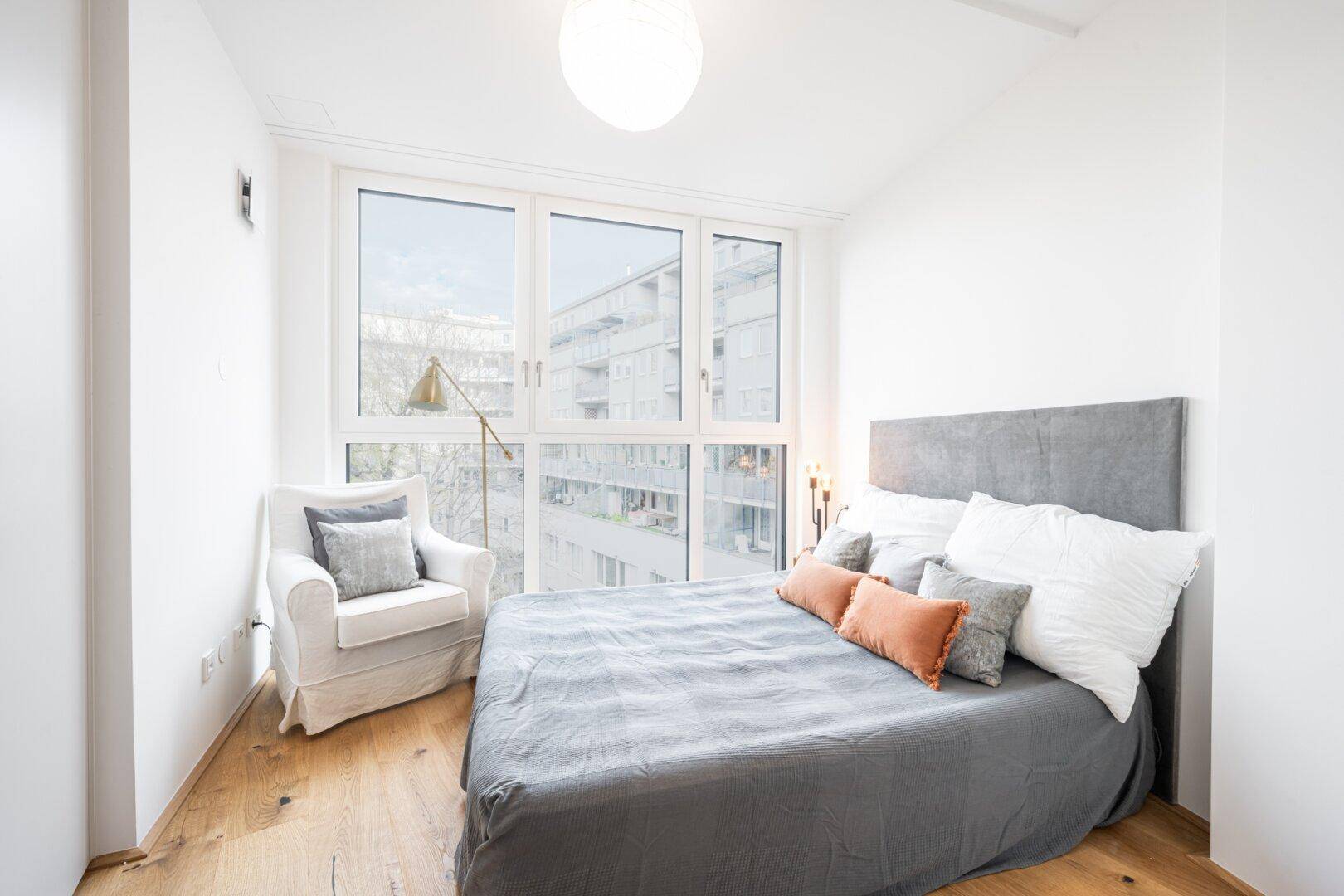 Helles Schlafzimmer mit großem Fenster, Holzboden und gemütlichem Sessel.