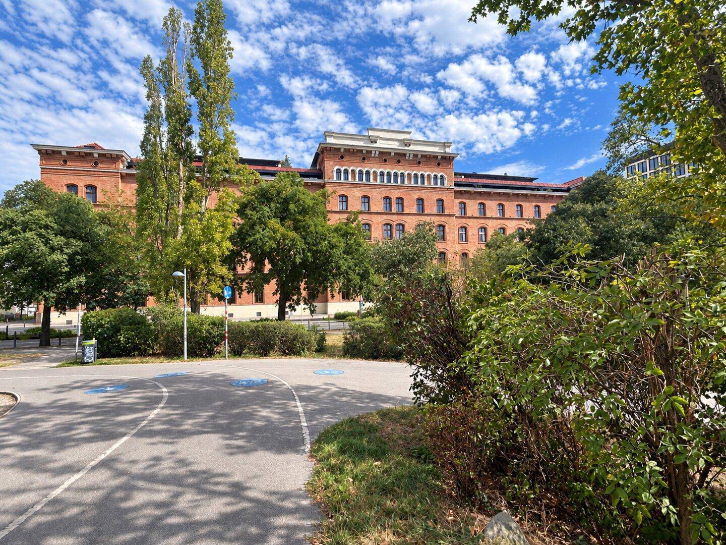 Historisches Backsteingebäude mit vielen Fenstern, umgeben von Bäumen und Grünflächen.