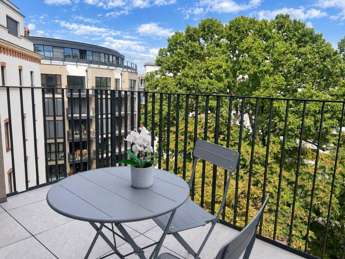 Sonniger Balkon mit Sitzgelegenheiten und Blick auf grüne Bäume und umliegende Gebäude.