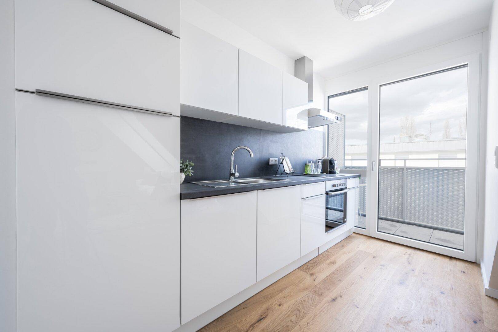 Moderne Einbauküche mit weißen Schränken, dunkler Arbeitsplatte und Zugang zum Balkon.