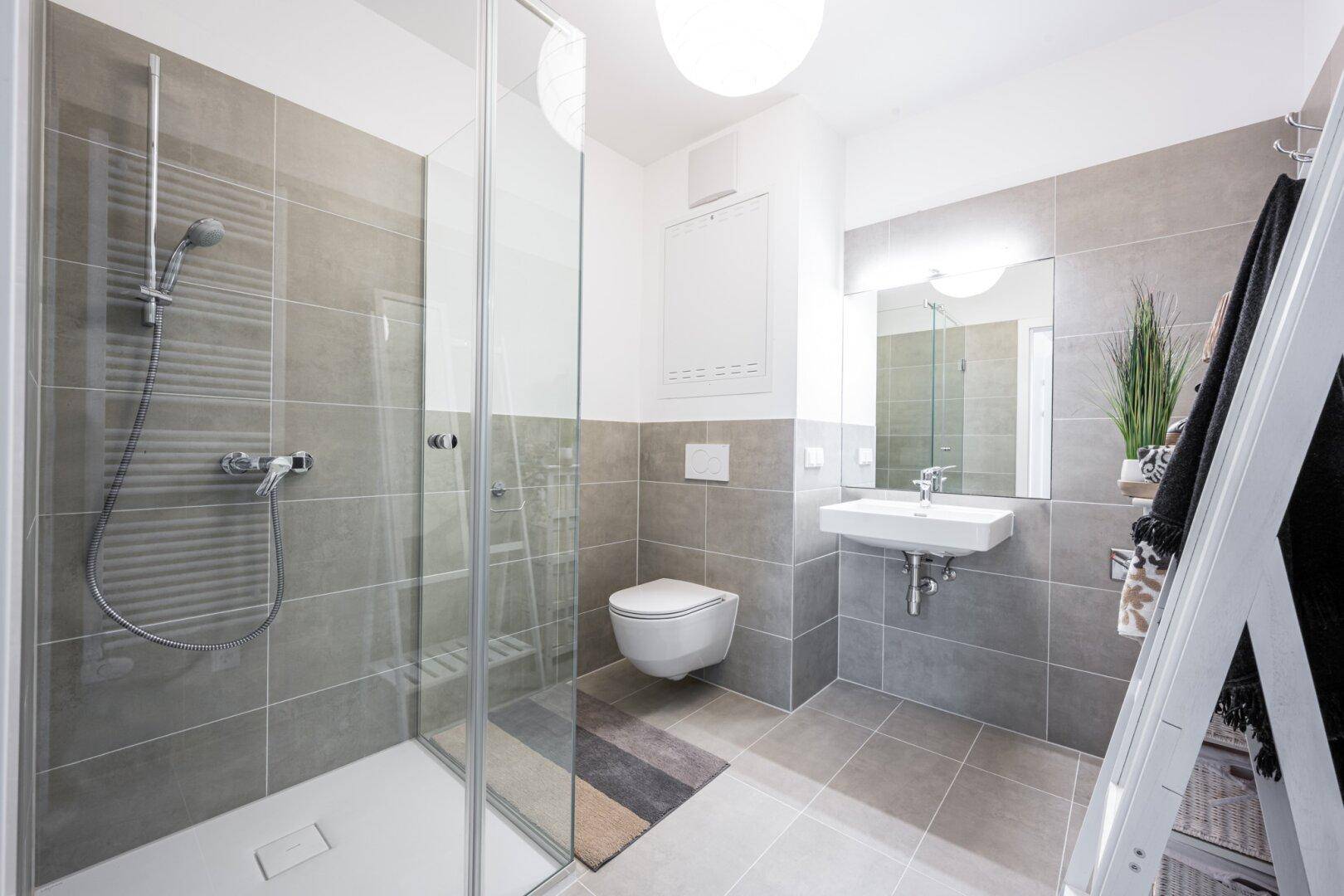 Helles Badezimmer mit begehbarer Dusche, Wand-WC und grauen Fliesen.