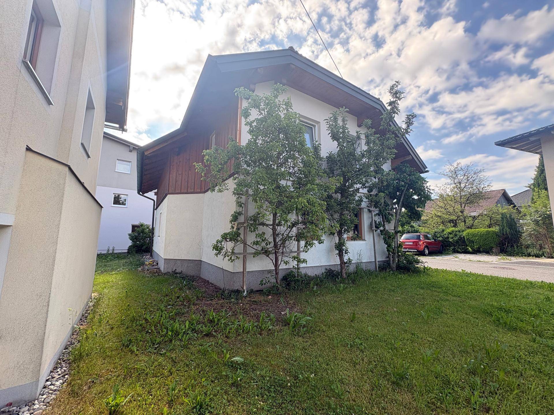Seitenansicht des Hauses mit einer grünen Rasenfläche und jungen Bäumen unter bewölktem Himmel.