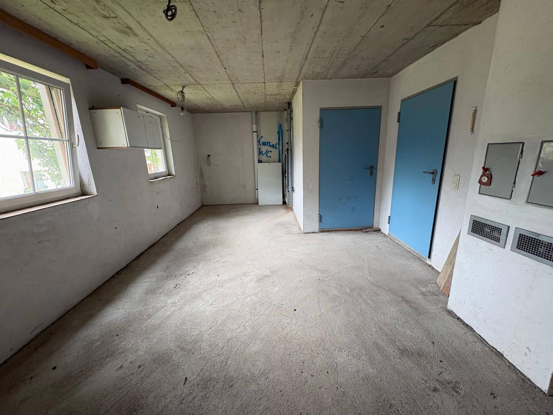 Funktionaler Raum mit Betonboden, zwei blauen Türen und einem Fenster mit Schrank.