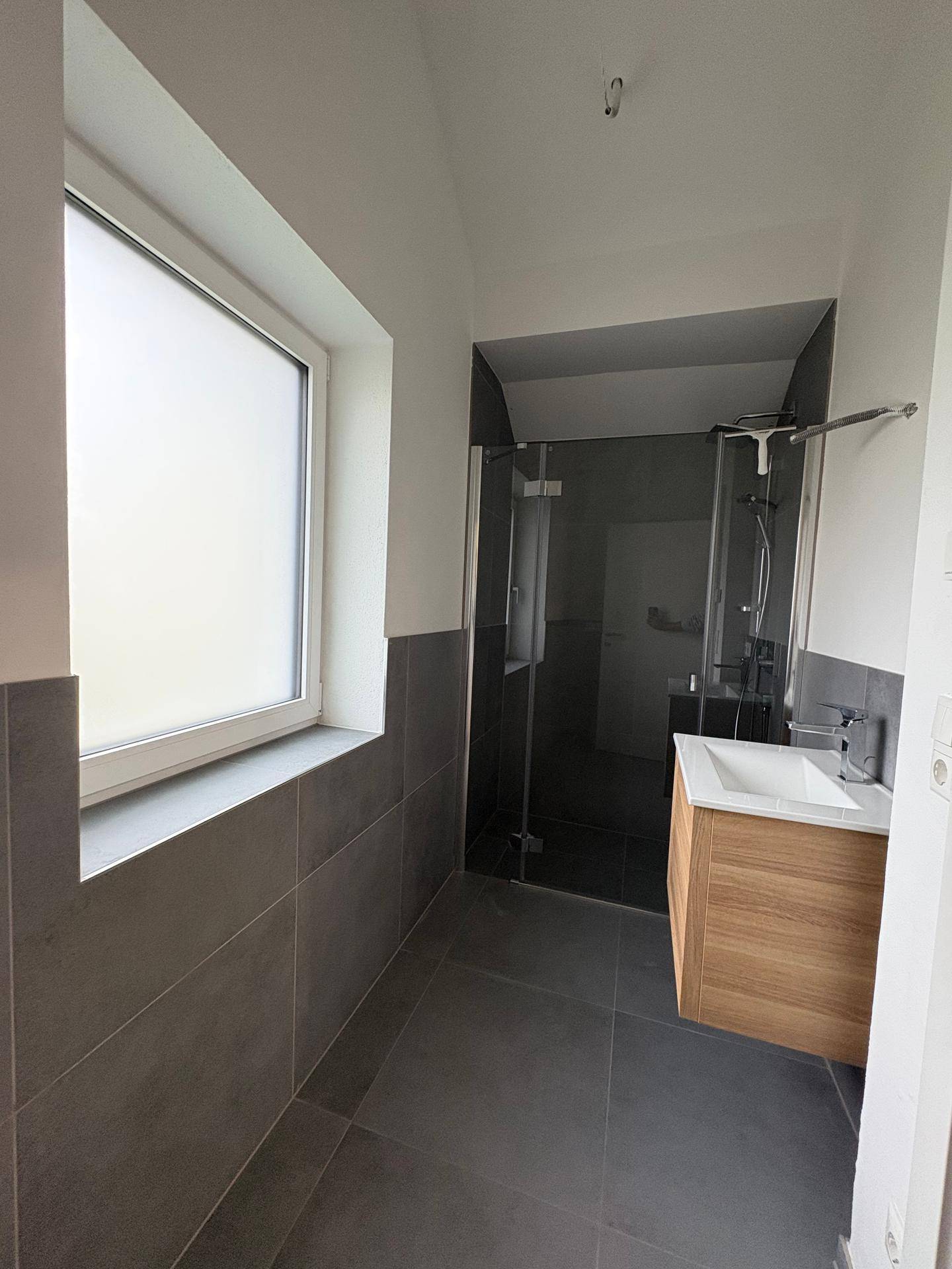 Modernes Badezimmer mit großer Dusche, Holzwaschtisch, grauen Fliesen und Fenster.