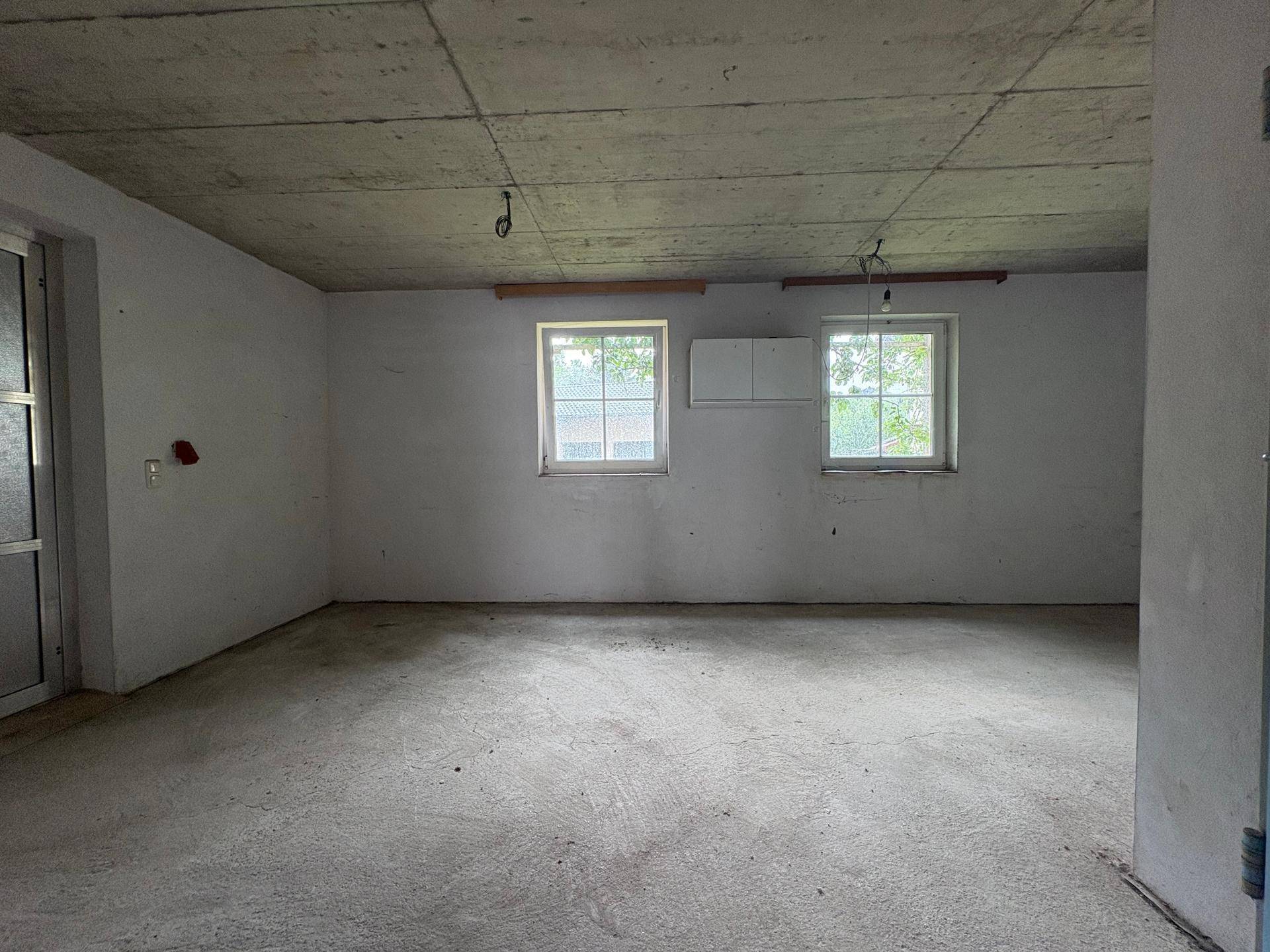 Leerer Raum mit Betonboden, zwei Fenstern und unverputzter Betondecke.