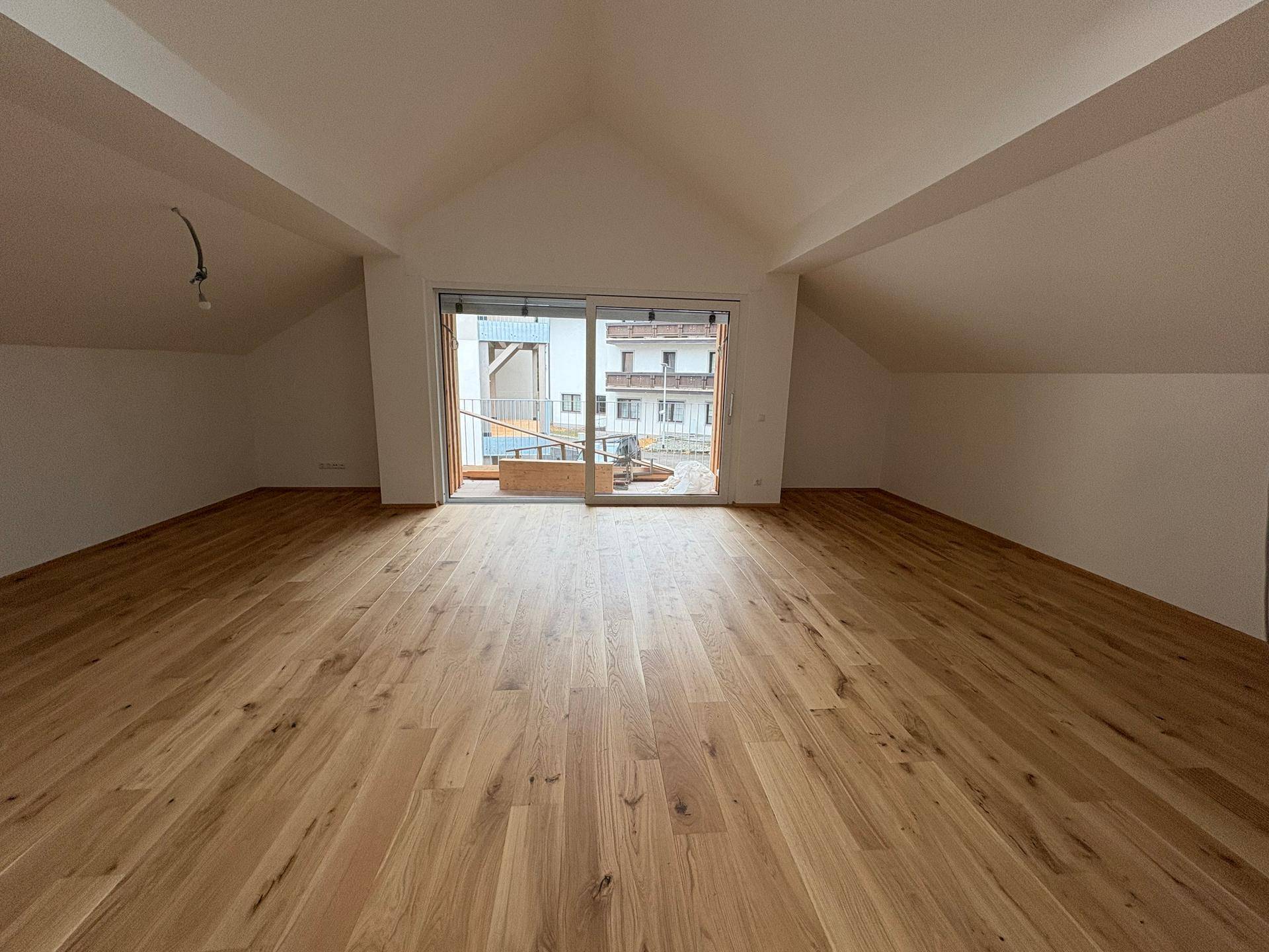 Geräumiger Wohnbereich im Dachgeschoss mit Holzboden und Zugang zum Balkon.
