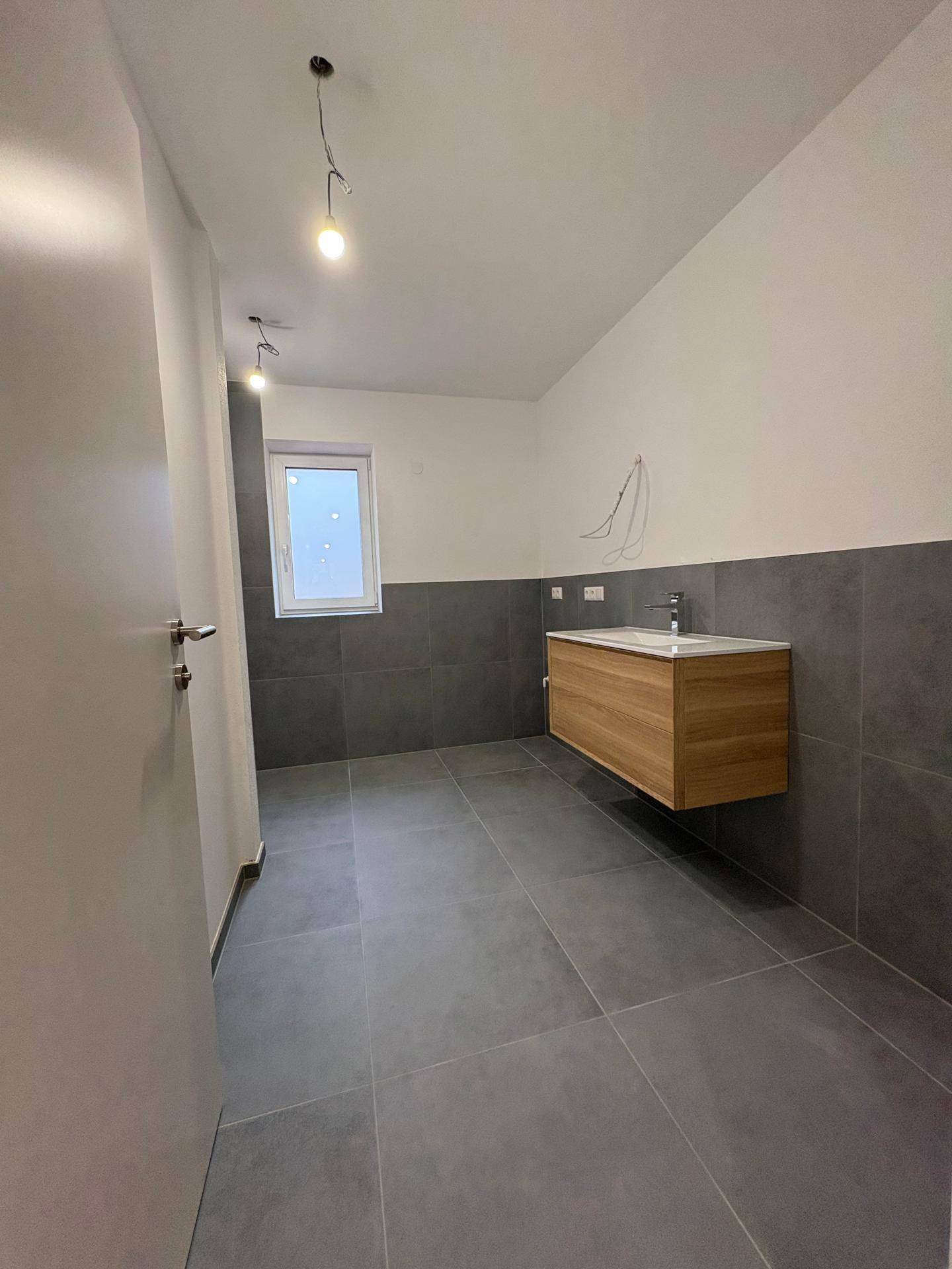 Modernes Badezimmer mit grauen Fliesen, schwebendem Waschtisch und Fenster.