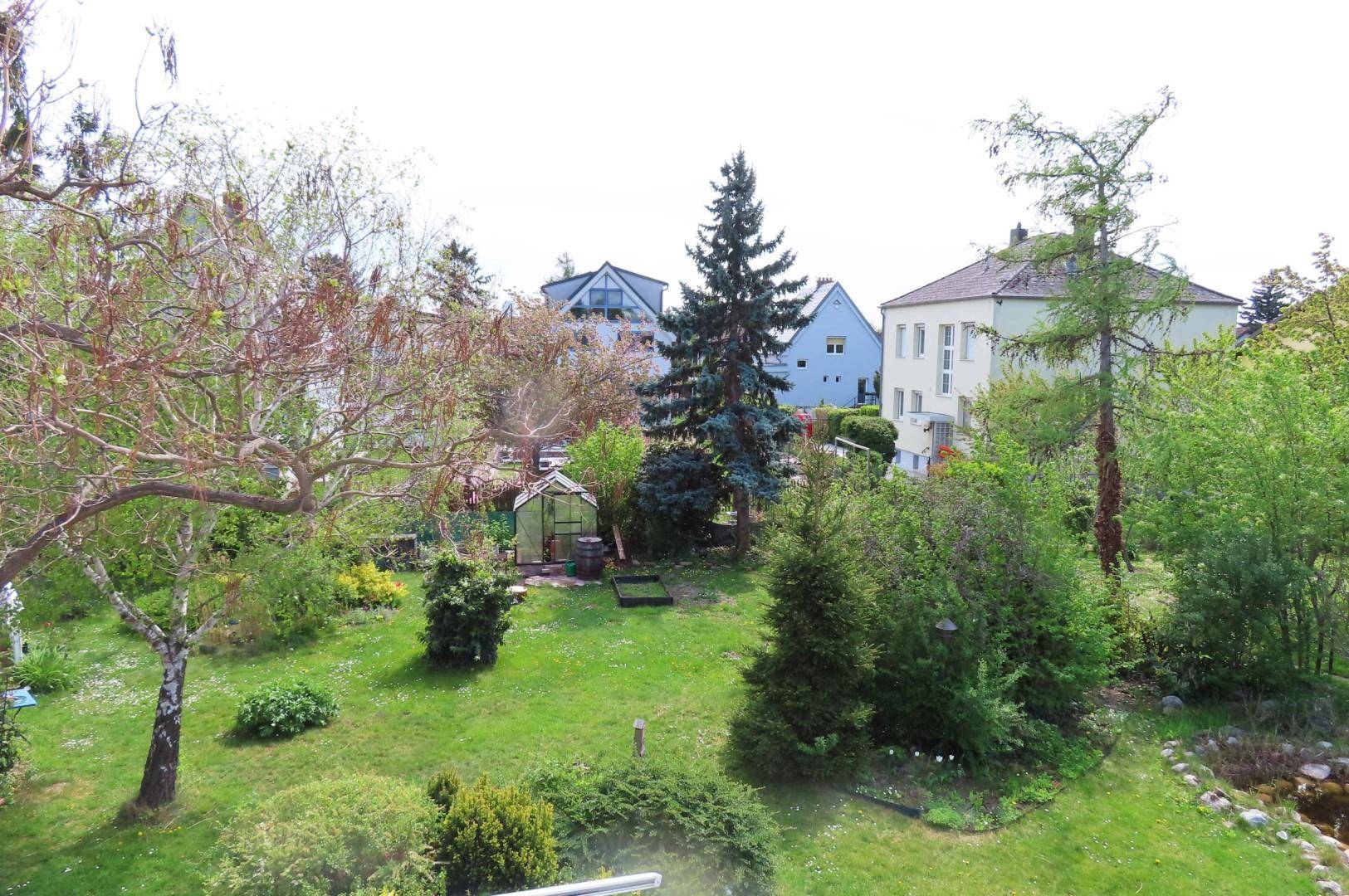 Weitläufiger Gartenbereich von oben mit einem kleinen Gewächshaus, Bäumen und umliegenden Häusern.