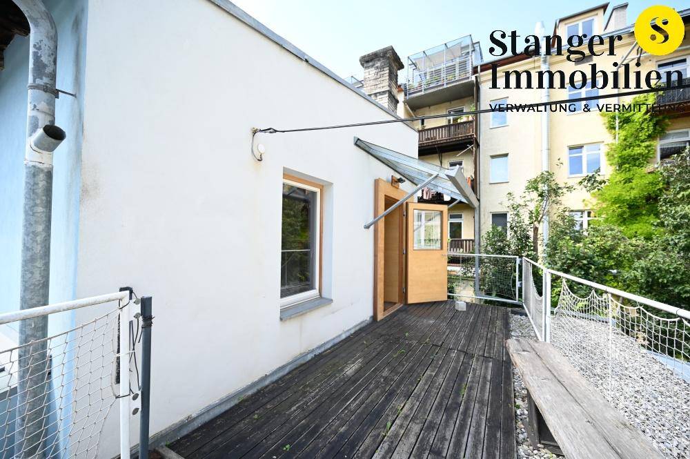 Helle Holzterrasse mit Sitzbank und Zugang zum Gebäude, umgeben von Grünflächen.