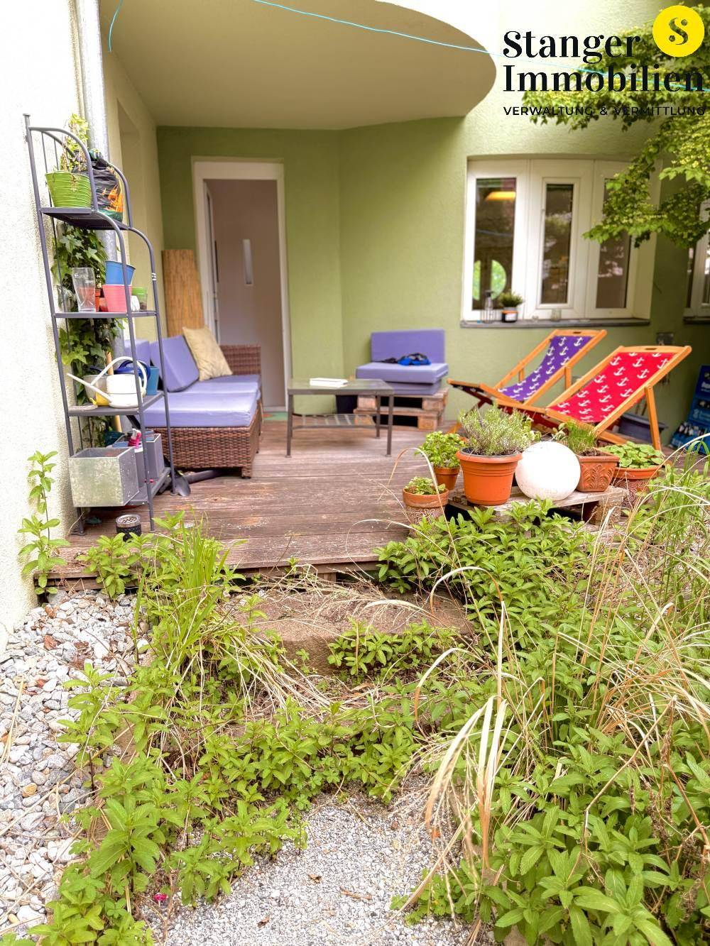 Gemütliche Terrasse mit Holzboden und Sitzgelegenheiten, umgeben von Grünpflanzen und Kies.