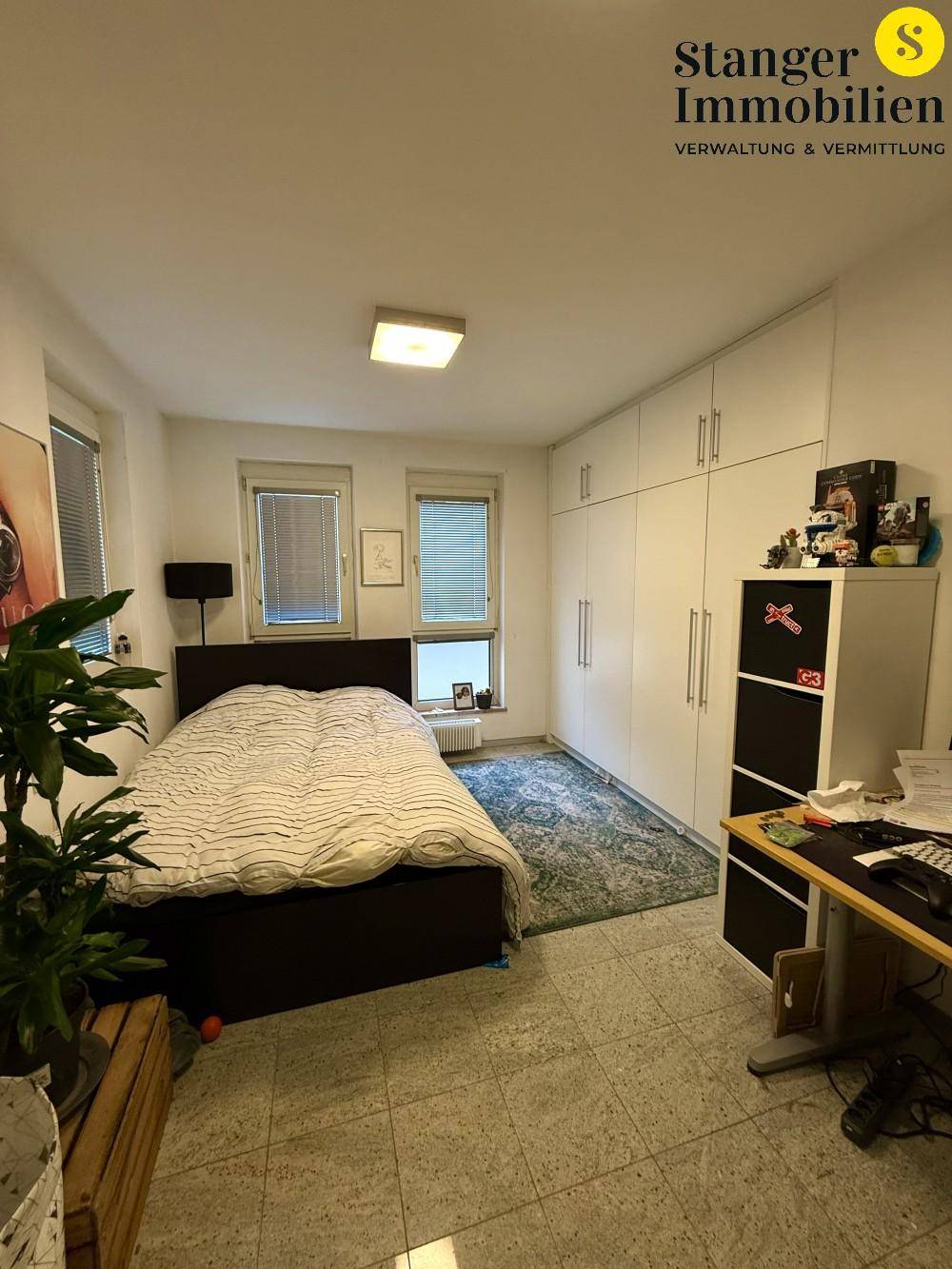 Helles Schlafzimmer mit großem Bett, Einbauschränken und zwei Fenstern, auf Granitfliesenboden.