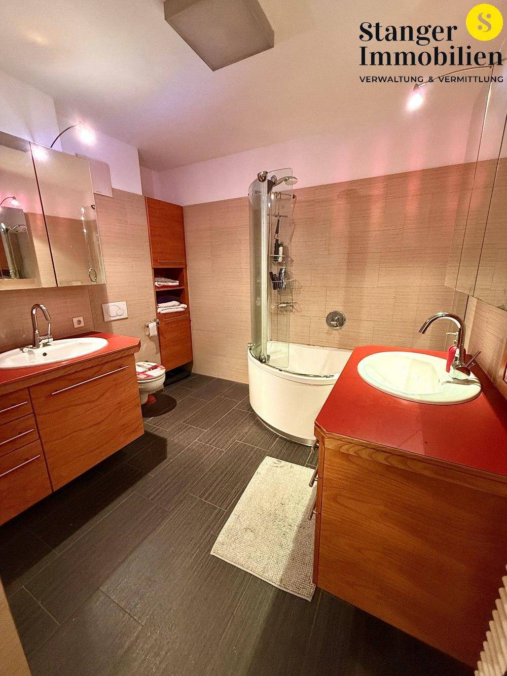 Badezimmer mit zwei Waschbecken, einer Duschkabine und dunklen Fliesen, modern eingerichtet.
