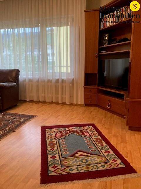 Wohnzimmer mit großem Holzschrank, Fernseher und Fenster mit Vorhängen für Privatsphäre.