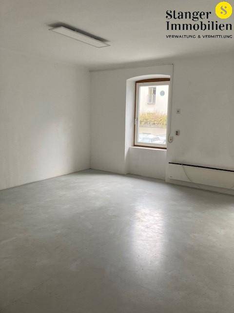 Heller, leerer Raum mit grauem Boden, einem Fenster und Heizkörpern unter der Brüstung.