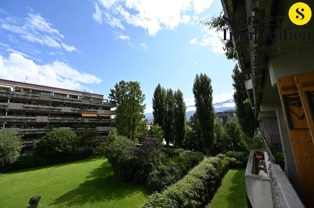 Weitläufiger Blick vom Balkon auf eine gepflegte Grünanlage und hohe Bäume unter blauem Himmel.