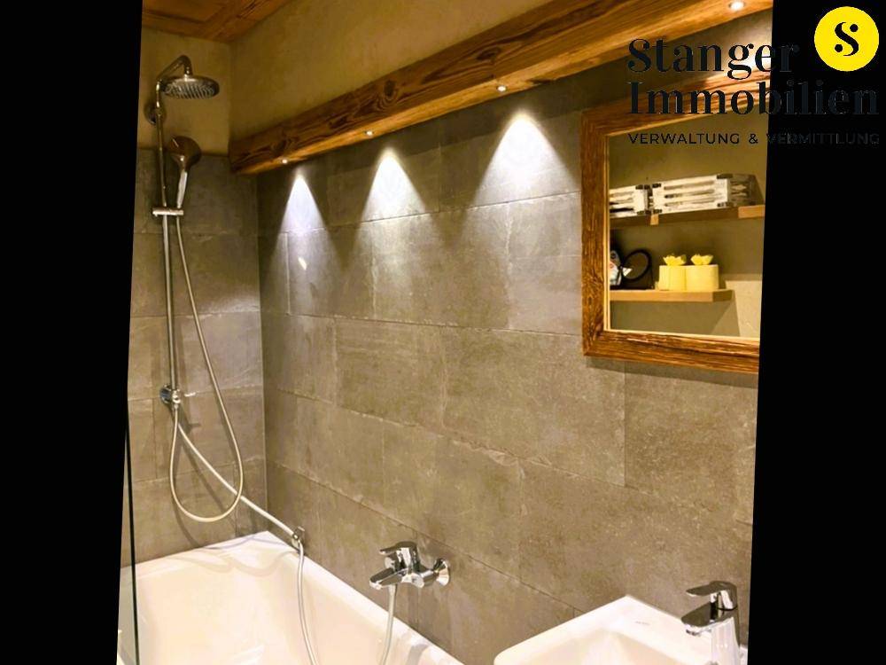 Duschbereich im Badezimmer mit Badewanne, grauen Fliesen und indirekter Beleuchtung durch Holzdetails.
