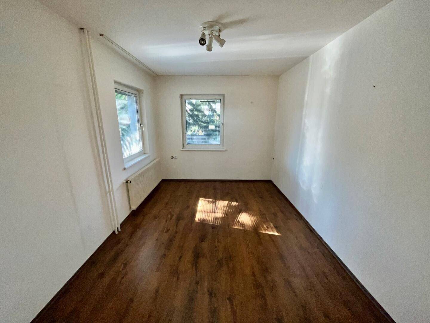 Gemütliches Schlafzimmer mit dunklem Holzfußboden und zwei Fenstern, die viel Licht hereinlassen.