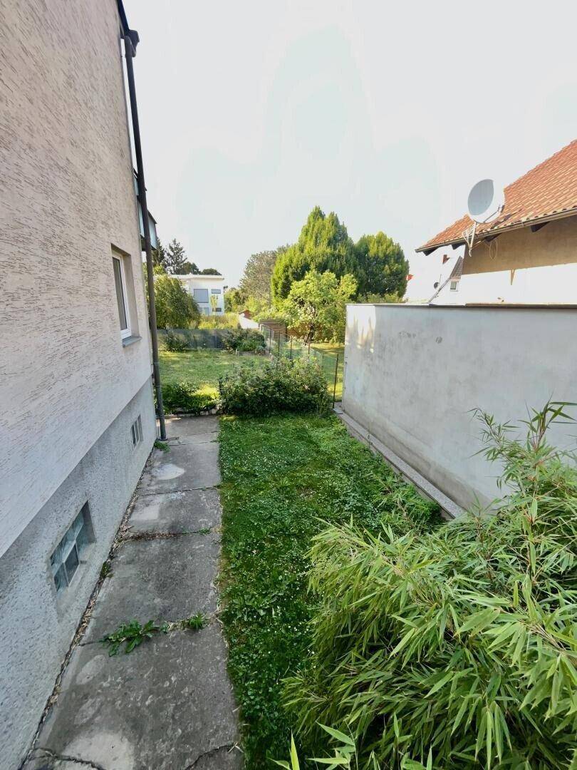 Schmaler Gartenweg entlang der Hauswand mit grüner Bepflanzung und Betonplatten.