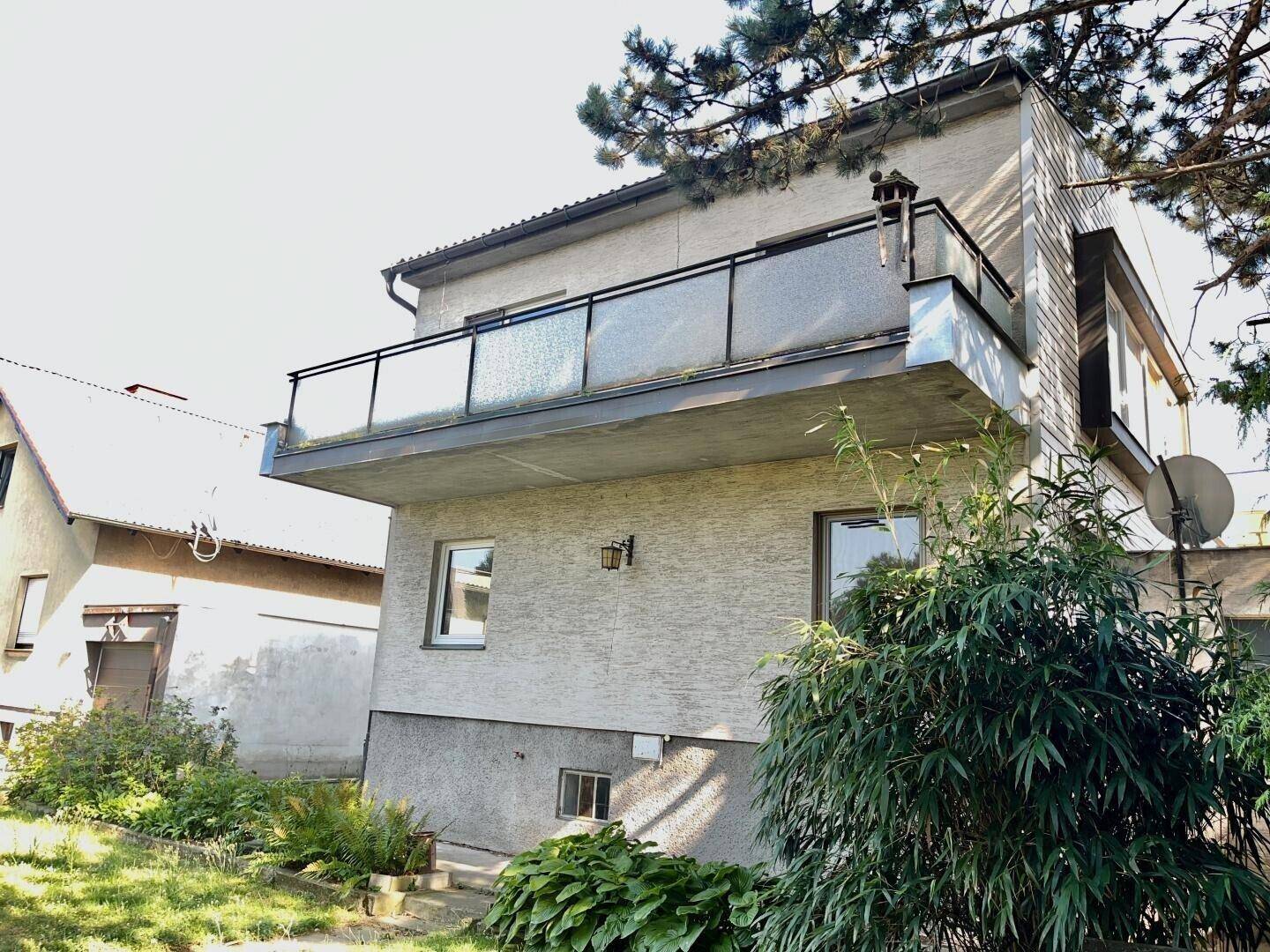 Nahaufnahme der Hausfassade mit Balkon und Glasgeländer, umgeben von üppigem Grün.