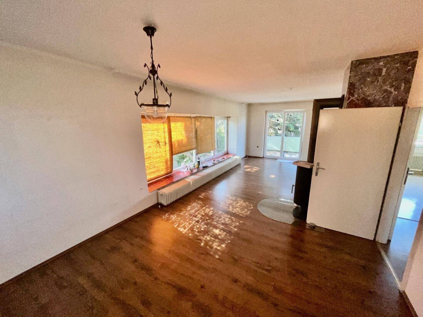 Geräumiges Wohnzimmer mit Kaminofen, Holzboden und großen Fenstern mit Blick ins Grüne.