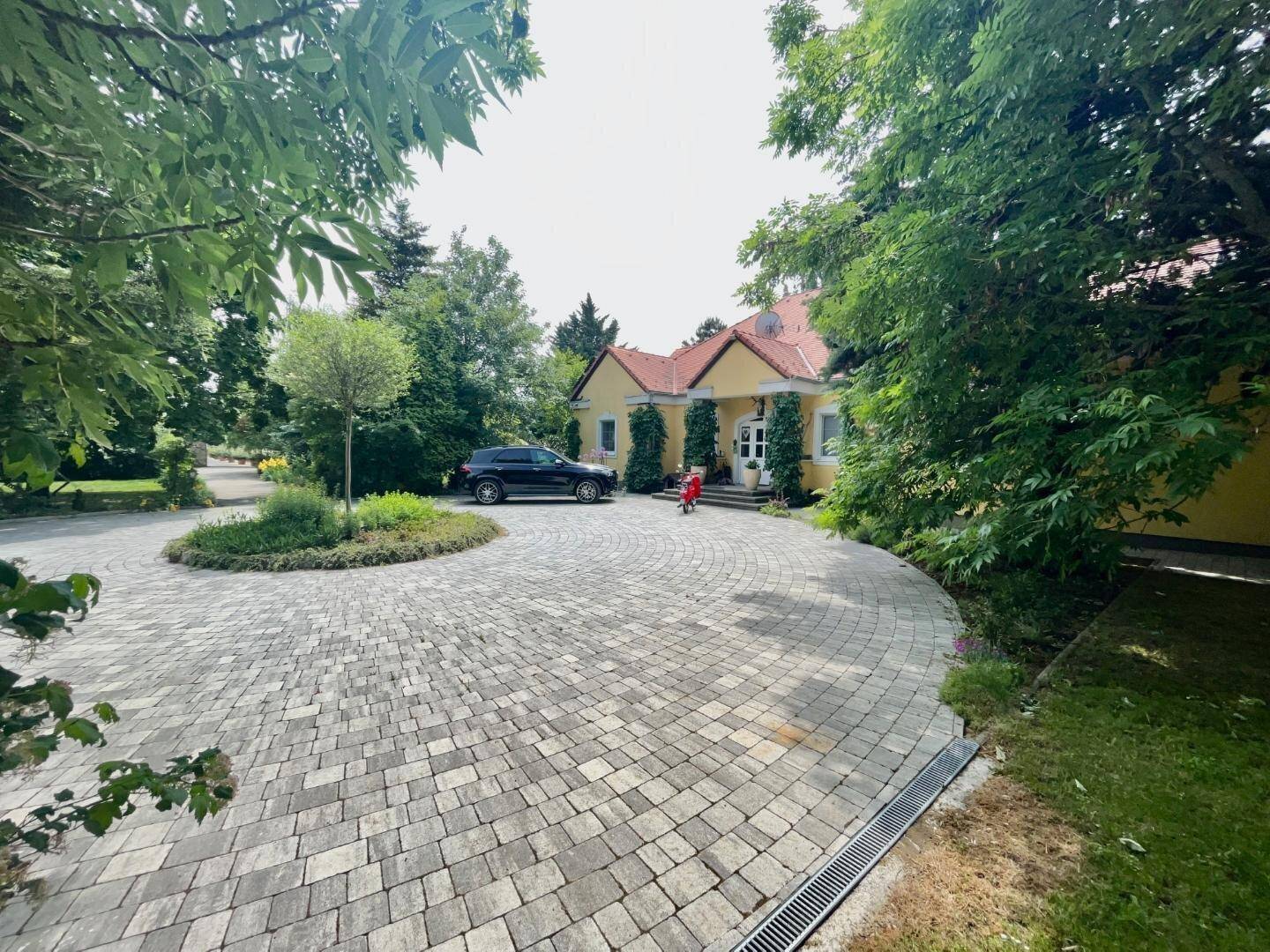 Eingangsbereich des Hauses mit gepflasterter Auffahrt, umgeben von Bäumen und Grünflächen.