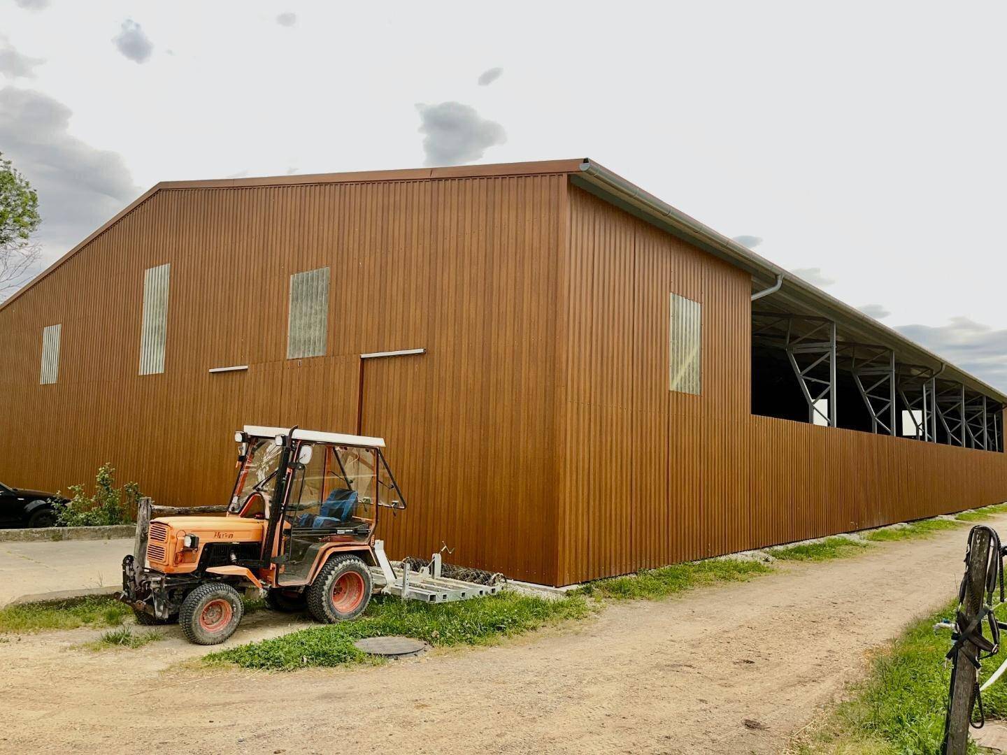 Modernes Stallgebäude mit brauner Holzverkleidung und einem Traktor davor unter bewölktem Himmel.
