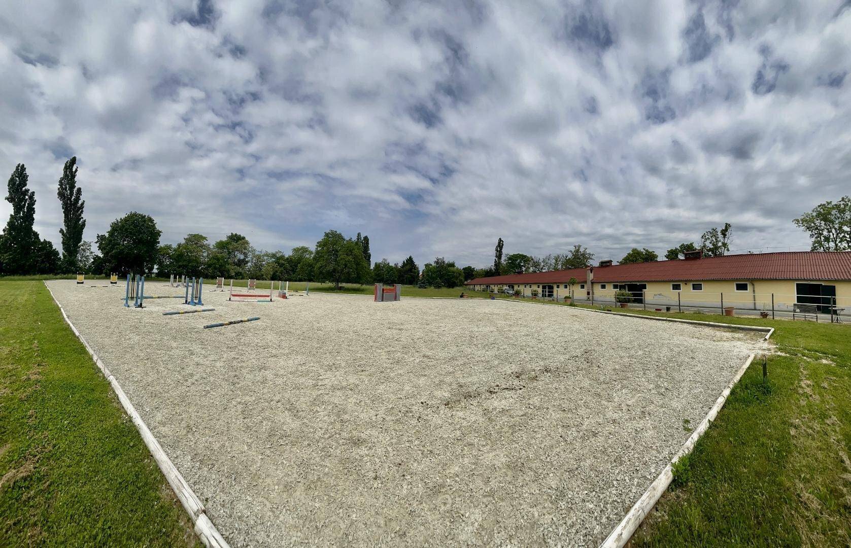 Großer Reitplatz im Freien mit Sandboden und Hindernissen, umgeben von Gebäuden und Bäumen.