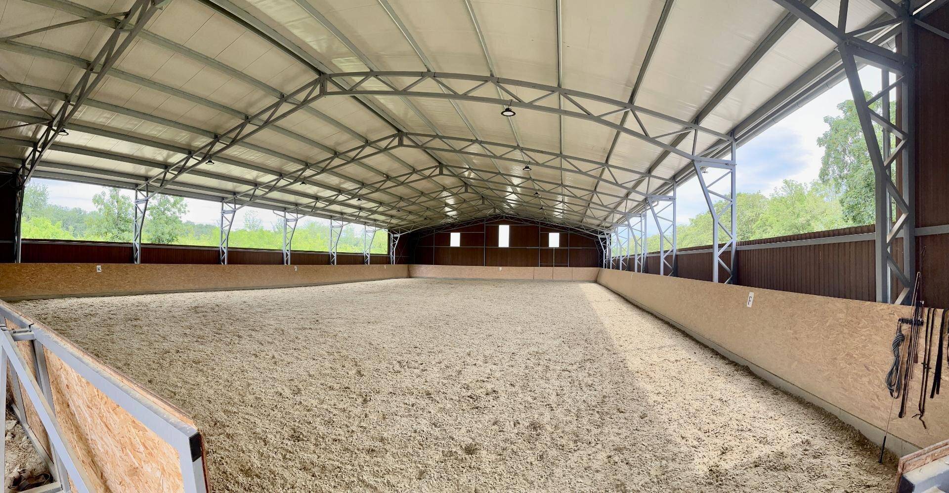 Große helle Reithalle mit Sandboden, Metallkonstruktion und viel Tageslicht durch offene Seiten.